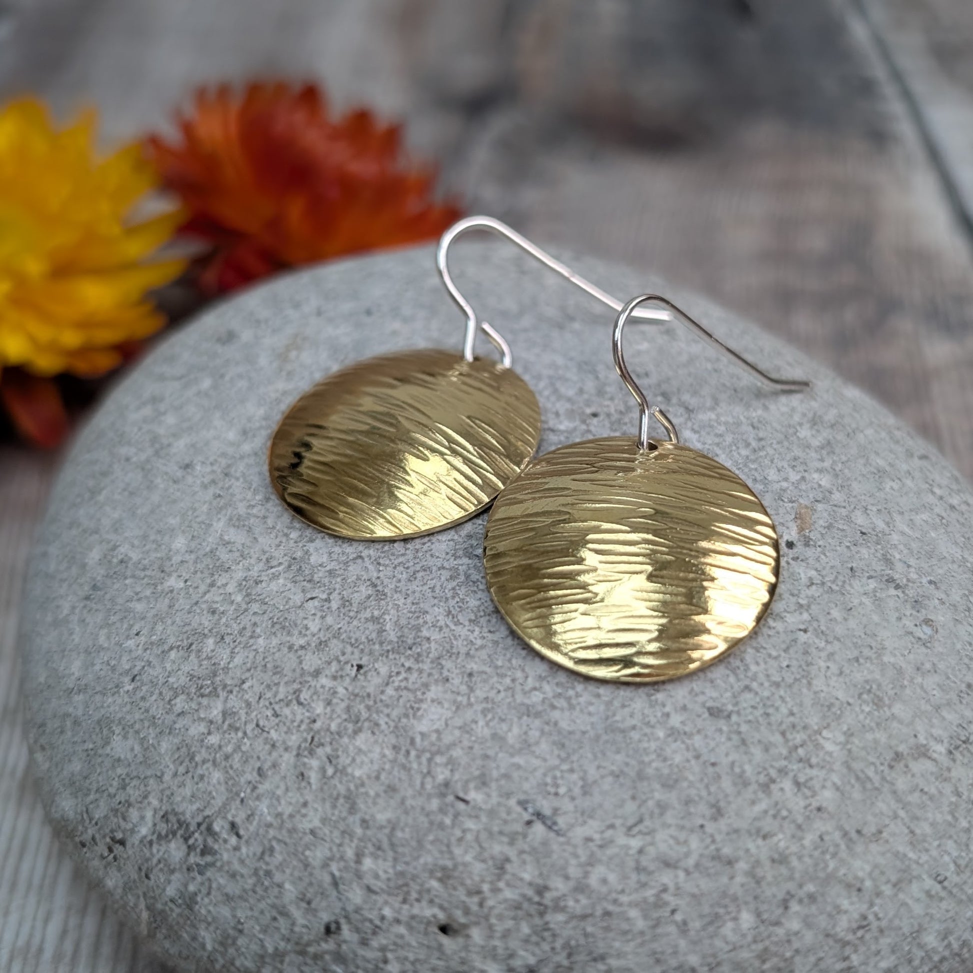 Two hammered brass domed disc earrings resting on a smooth grey pebble, with sterling silver hooks and autumn-coloured flowers in the background.