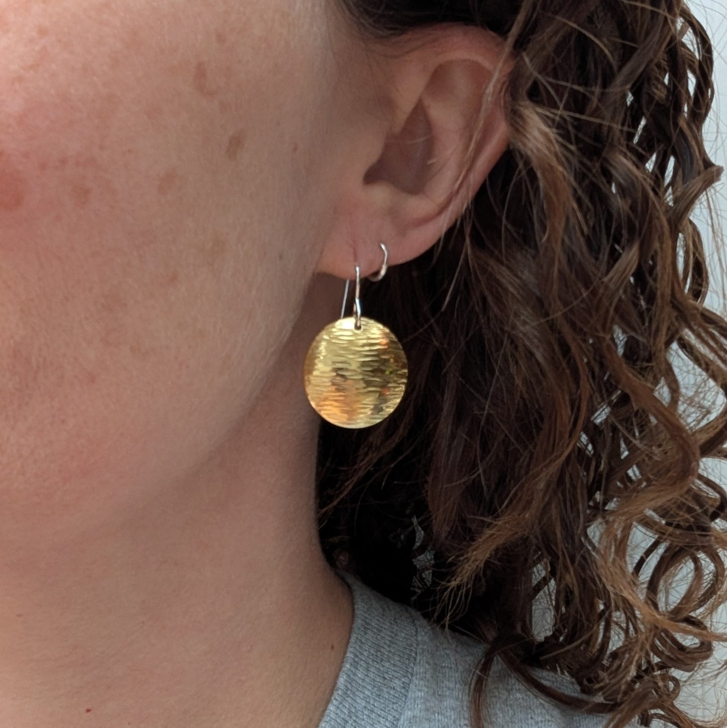 A person wearing one of the hammered brass domed disc earrings, showing its size and gentle dome shape against curly brown hair.