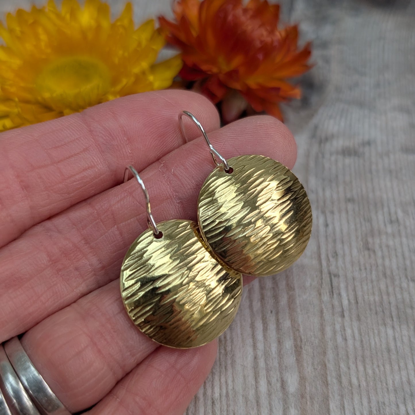 A pair of brass domed disc earrings held in a hand, showing the hammered texture and sterling silver ear hooks.