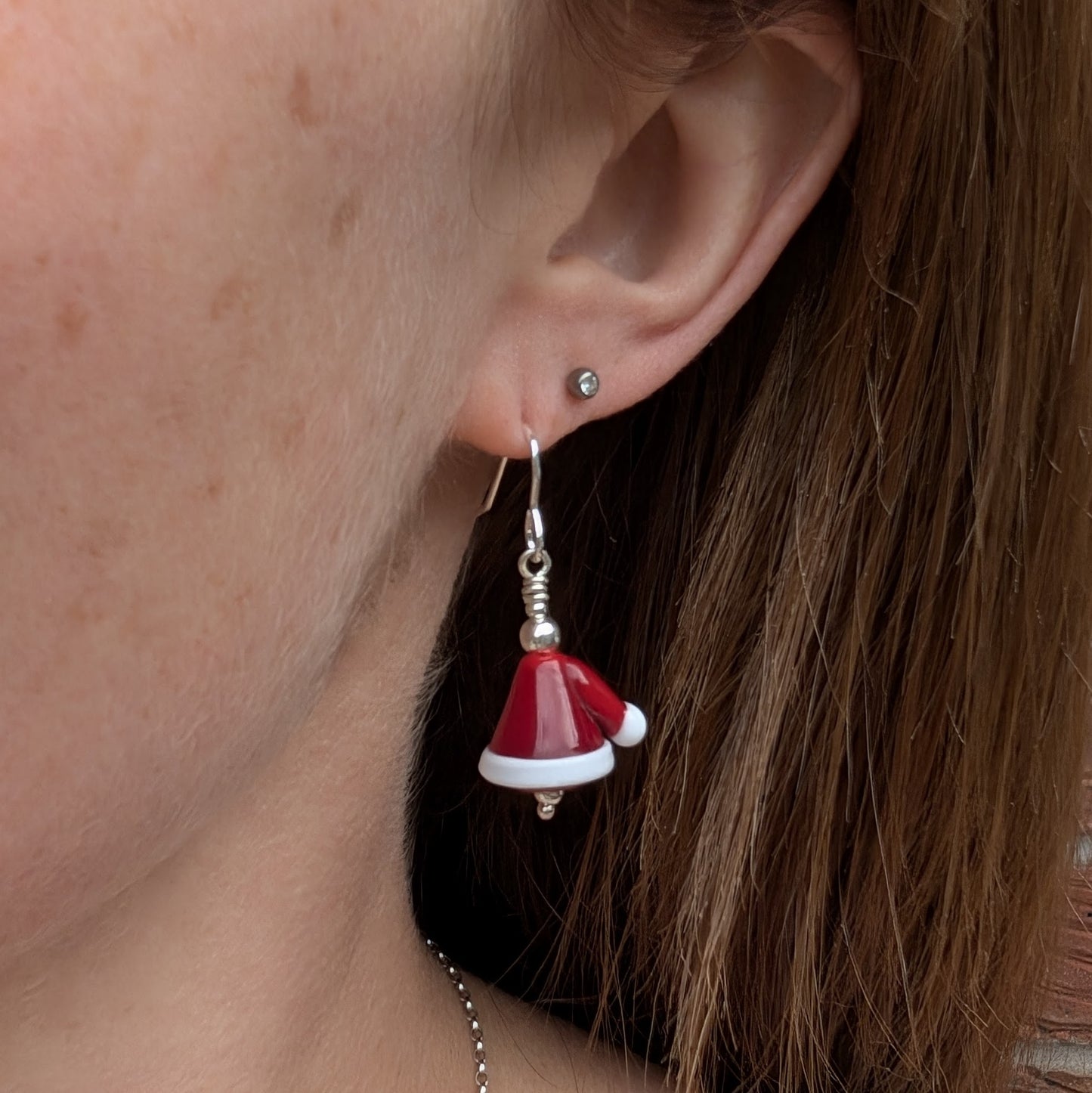 Each earring is a Santa hat-shape made from red and white lampwork glass. They are red with white trim and a small white pom-pom detail. Suspended from silver earring wire.