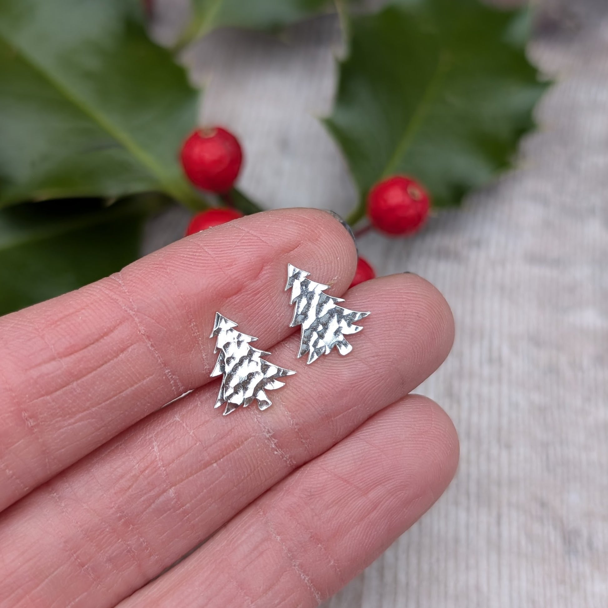 Close-up of a hand holding a pair of handmade silver Christmas tree stud earrings with a hammered texture, set against a background with holly leaves and red berries.