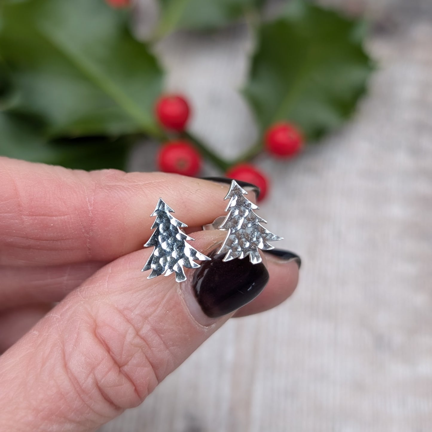 Close-up of a hand holding a pair of handmade silver Christmas tree stud earrings with a hammered texture, set against a background with holly leaves and red berries.