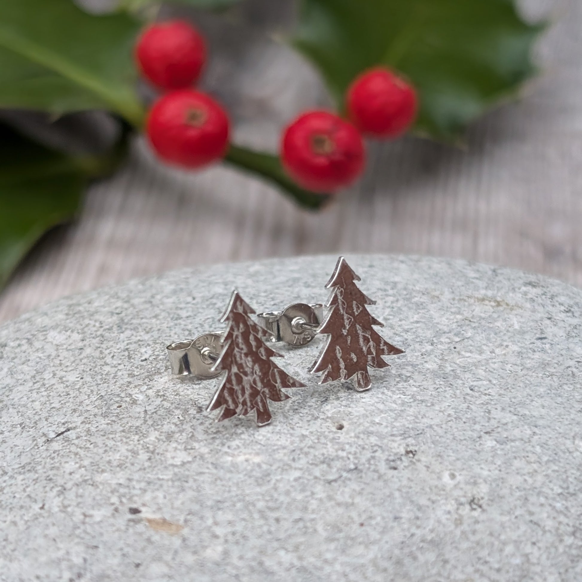 A pair of handmade silver Christmas tree stud earrings with a hammered texture resting on a grey stone surface, with a blurred background featuring holly leaves and red berries.