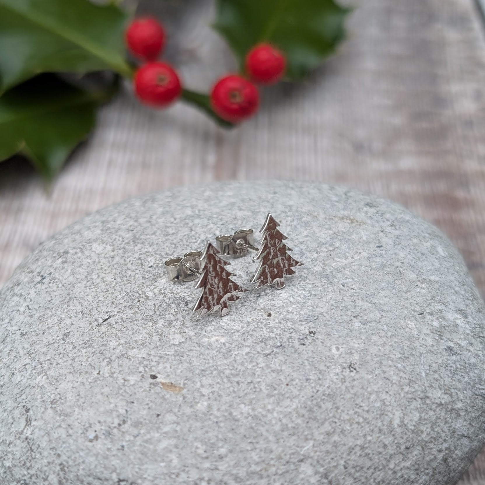 A pair of handmade silver Christmas tree stud earrings with a hammered texture resting on a grey stone surface, with a blurred background featuring holly leaves and red berries.