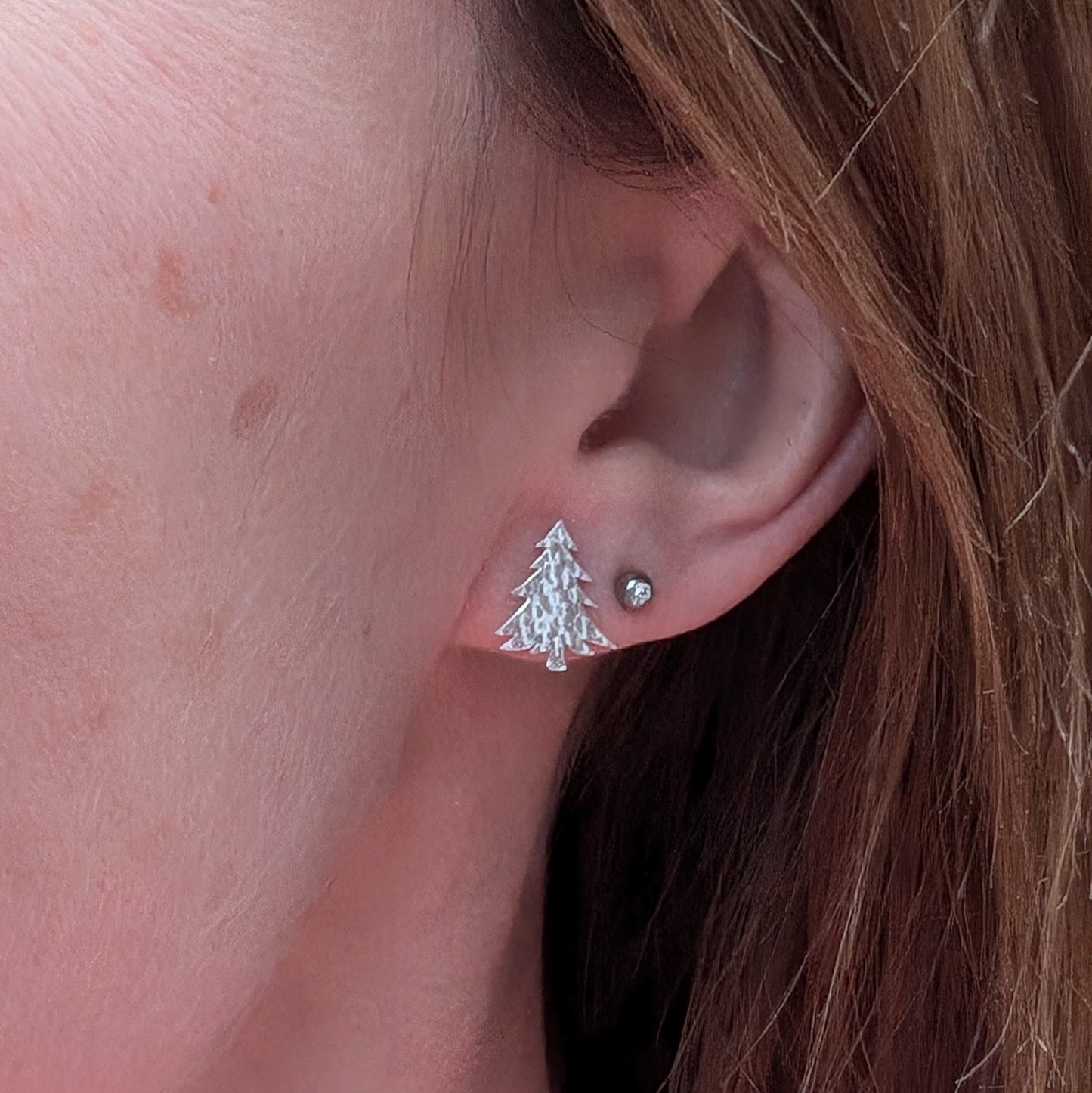 Close-up of a silver Christmas tree stud earring worn on an ear, showing its delicate hammered texture and festive shape.