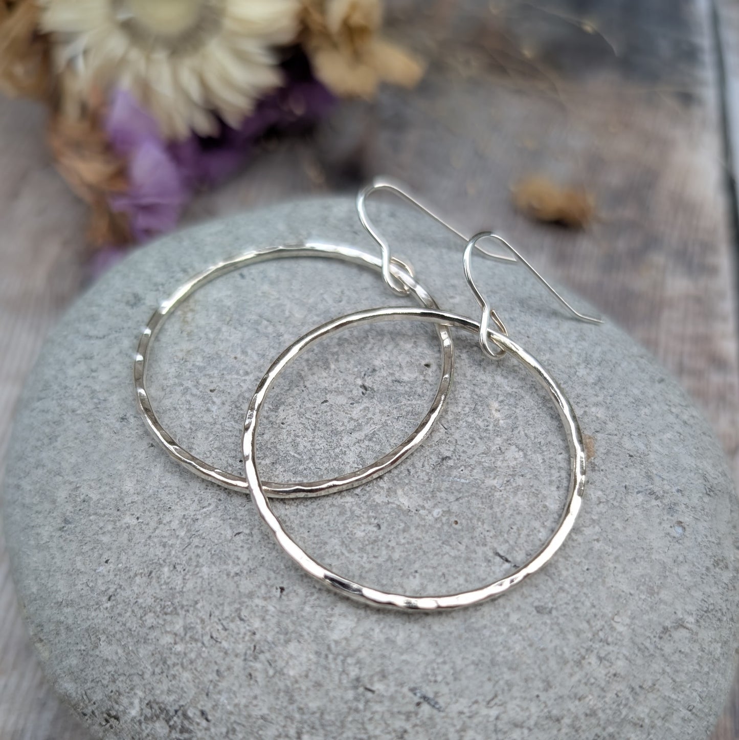 The hammered silver hoop earrings resting on a smooth grey stone. The earrings overlap slightly, showing their circular shape and textured surface. Dried flowers in the background provide a warm, neutral contrast.