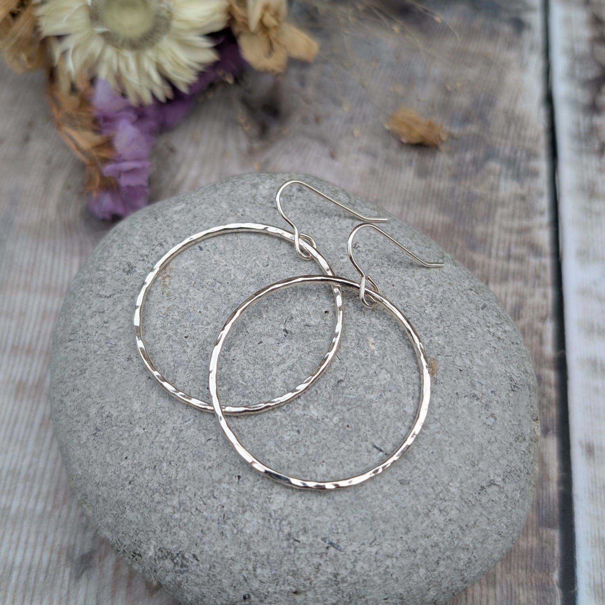 The hammered silver hoop earrings resting on a smooth grey stone. The earrings overlap slightly, showing their circular shape and textured surface. Dried flowers in the background provide a warm, neutral contrast.