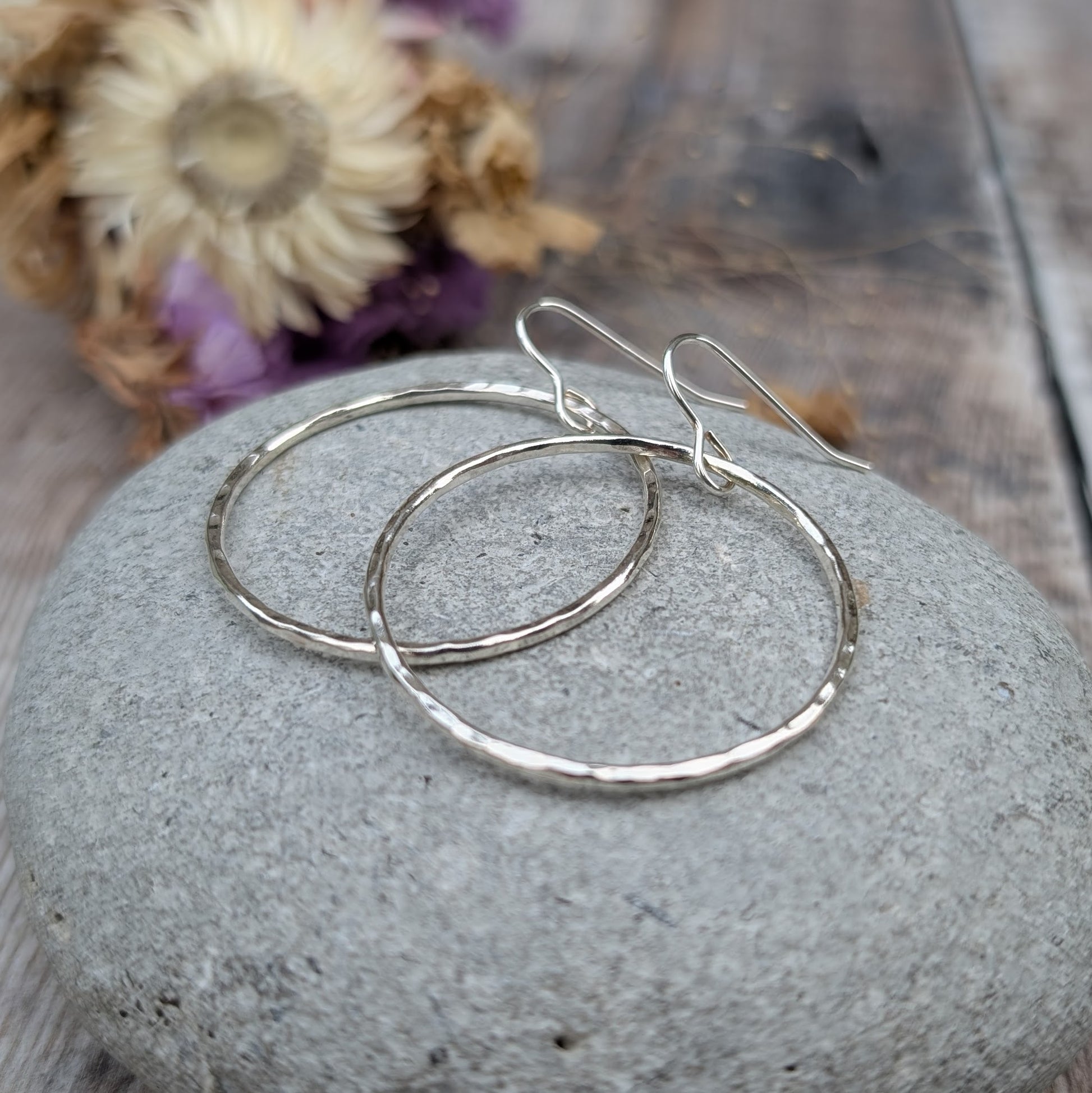 The hammered silver hoop earrings resting on a smooth grey stone. The earrings overlap slightly, showing their circular shape and textured surface. Dried flowers in the background provide a warm, neutral contrast.