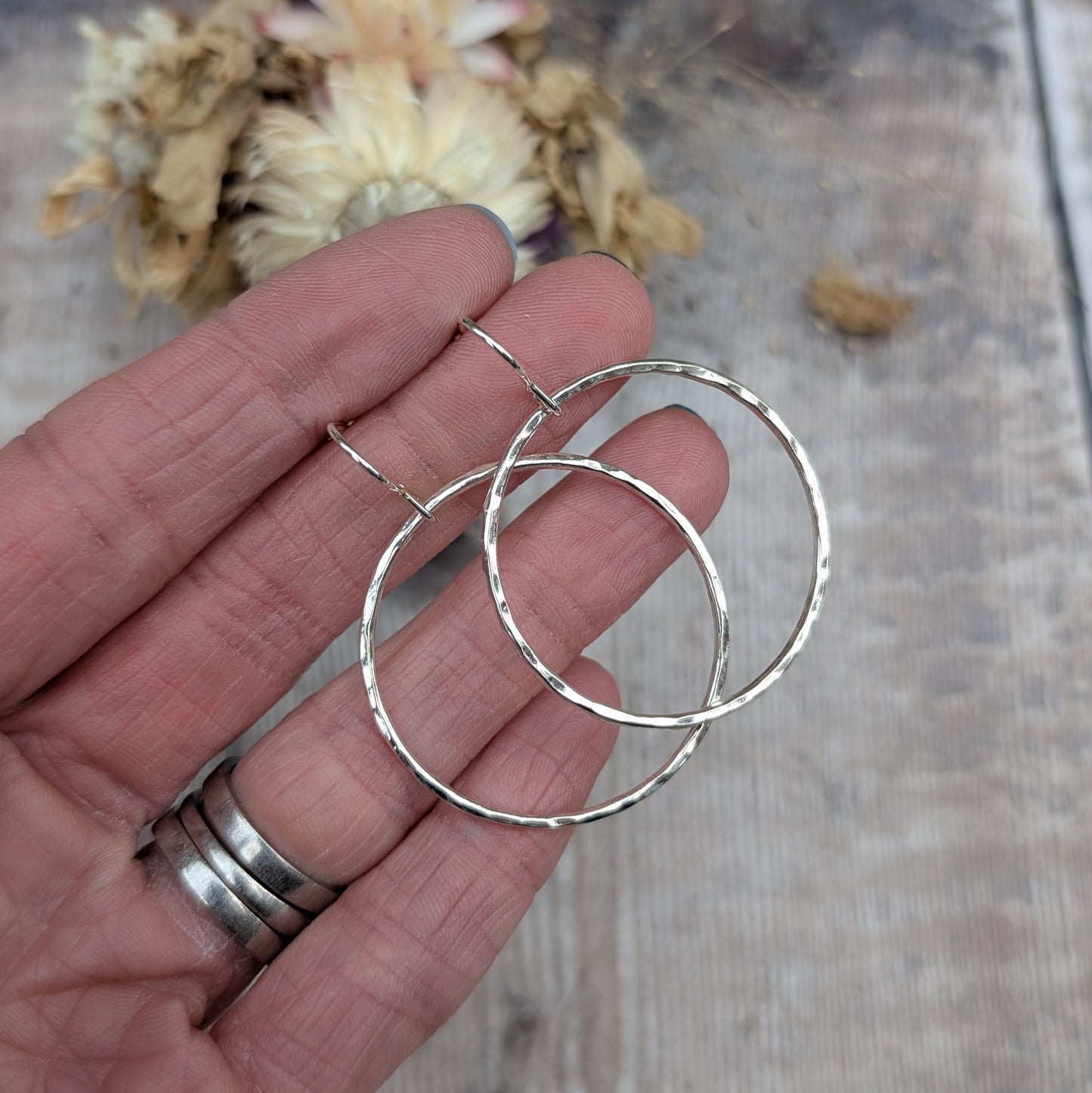 A pair of large handmade sterling silver hoop earrings with a hammered texture, held between fingers. The light catches the subtle facets, and dried flowers in the background add a soft, natural feel.