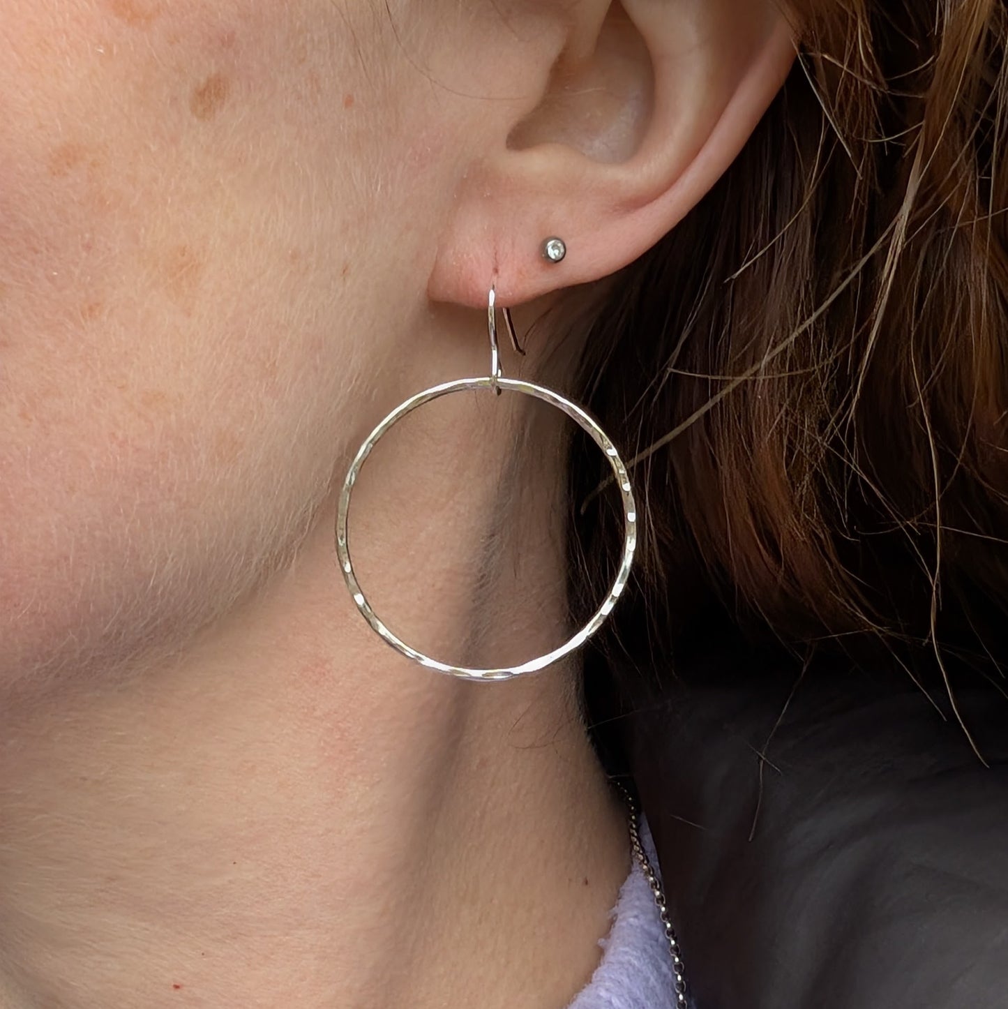 One hammered silver hoop earring being worn. The earring dangles just below the earlobe, its size and lightweight design making a statement. A small silver stud is also visible in the second piercing, with strands of hair framing the ear.