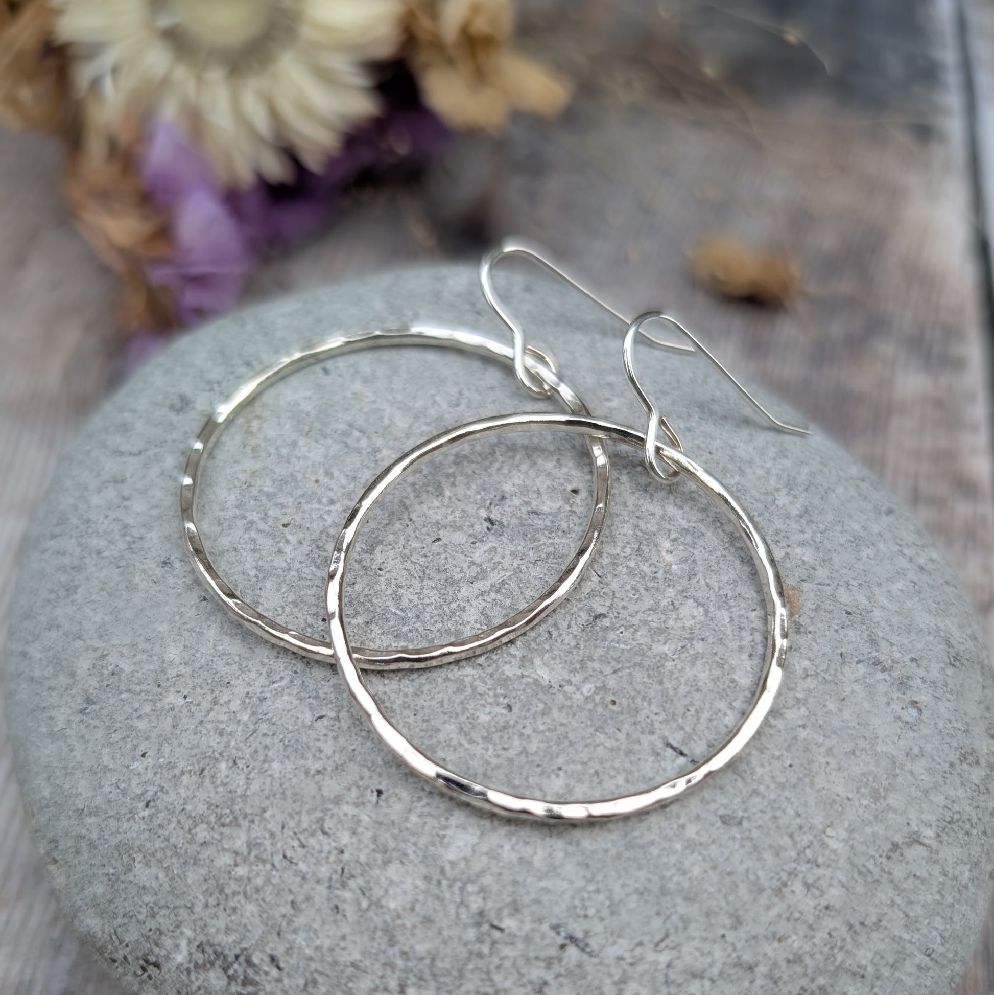 The hammered silver hoop earrings resting on a smooth grey stone. The earrings overlap slightly, showing their circular shape and textured surface. Dried flowers in the background provide a warm, neutral contrast.