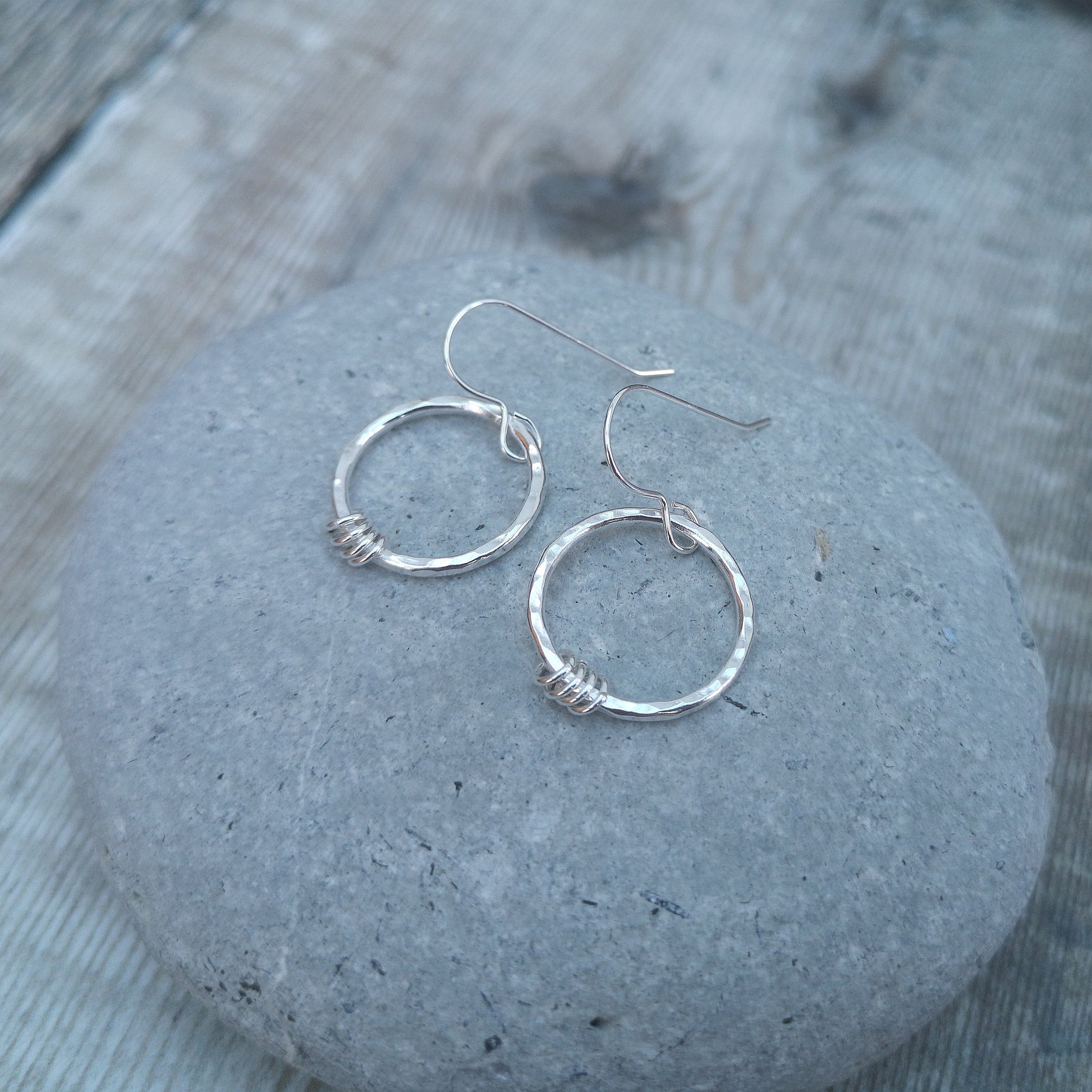 Close-up of sterling silver hammered circle earrings resting on a smooth grey stone. The earrings feature delicate silver loops wrapped around the base of the circles, showcasing their minimalist design and handmade craftsmanship.