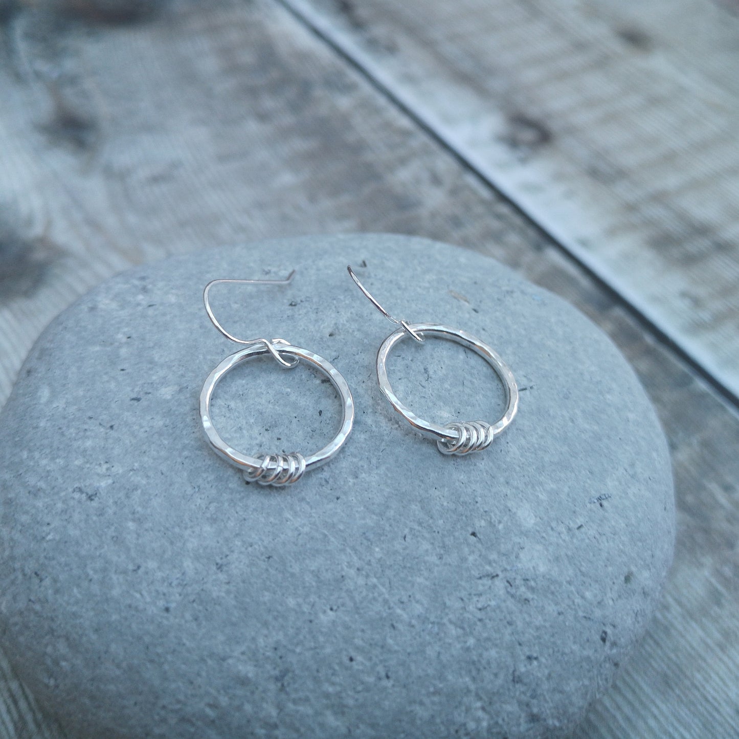 Close-up of sterling silver hammered circle earrings resting on a smooth grey stone. The earrings feature delicate silver loops wrapped around the base of the circles, showcasing their minimalist design and handmade craftsmanship.