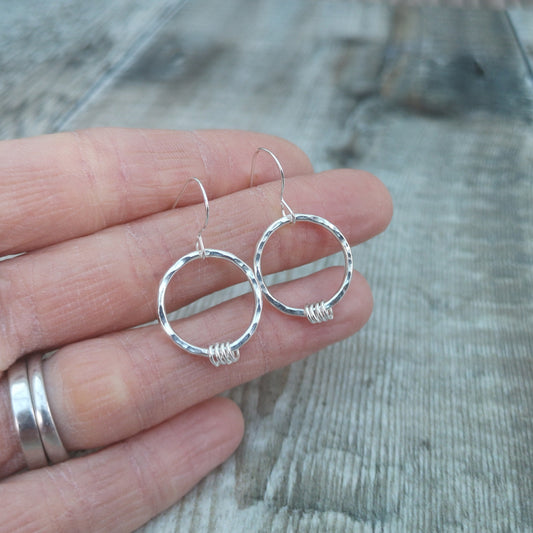 A hand holding a pair of sterling silver hammered circle earrings with wrapped silver loops at the base. The earrings reflect light, highlighting their textured surface and simple, elegant design.