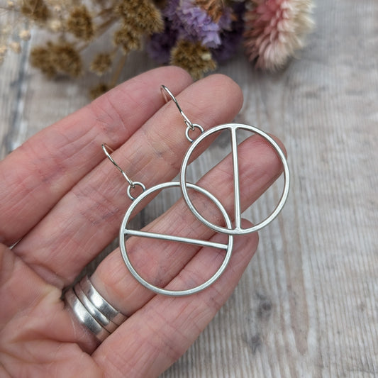 Close-up of a hand holding a pair of oxidised sterling silver circle earrings with a brushed satin finish, featuring a minimalist design with two diagonal lines across the center.