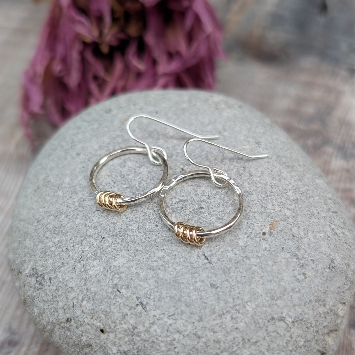 The silver circle earrings with gold loops resting on a smooth grey stone. The earrings overlap slightly, displaying their delicate yet sturdy design. Dried flowers in the background create a soft, natural setting.