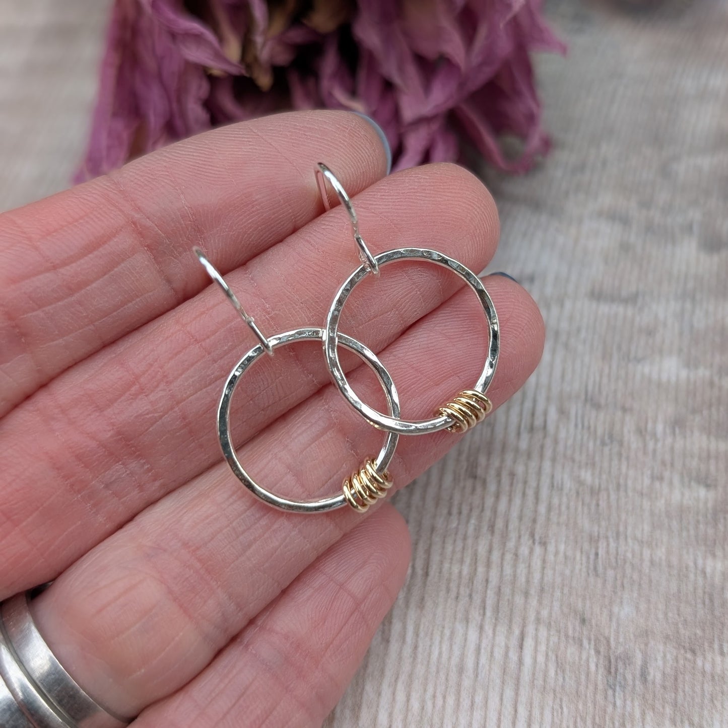 A pair of handmade sterling silver circle earrings with a hammered texture, each adorned with small gold loops. The earrings are held between fingers, showing their size and detailing, with dried purple flowers in the background.