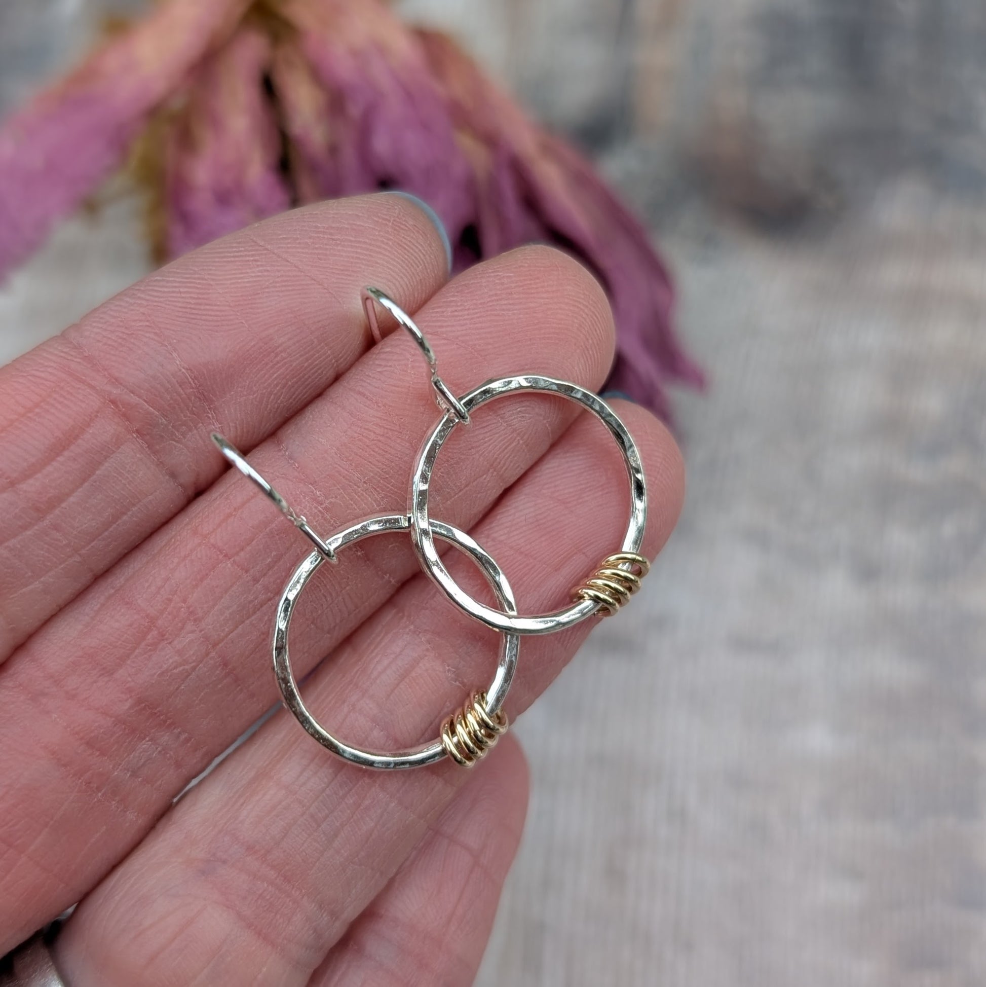 A pair of handmade sterling silver circle earrings with a hammered texture, each adorned with small gold loops. The earrings are held between fingers, showing their size and detailing, with dried purple flowers in the background.