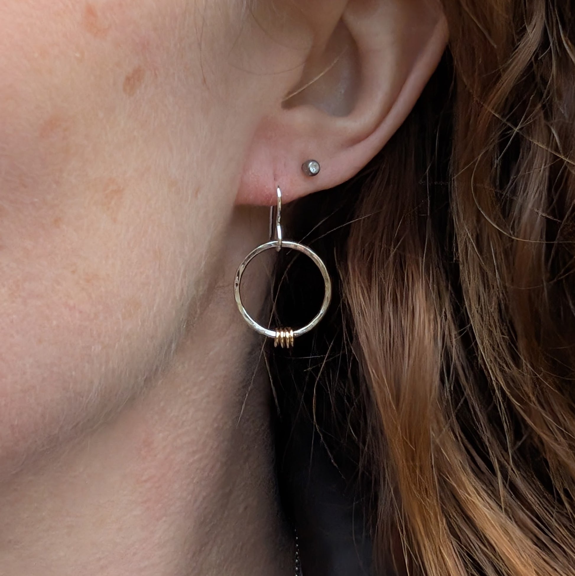 One silver and gold-accented circle earring being worn. The hammered silver hoop dangles just below the earlobe, with the small gold loops adding a warm contrast. A silver stud earring is visible in the second piercing, and strands of brown hair frame the ear