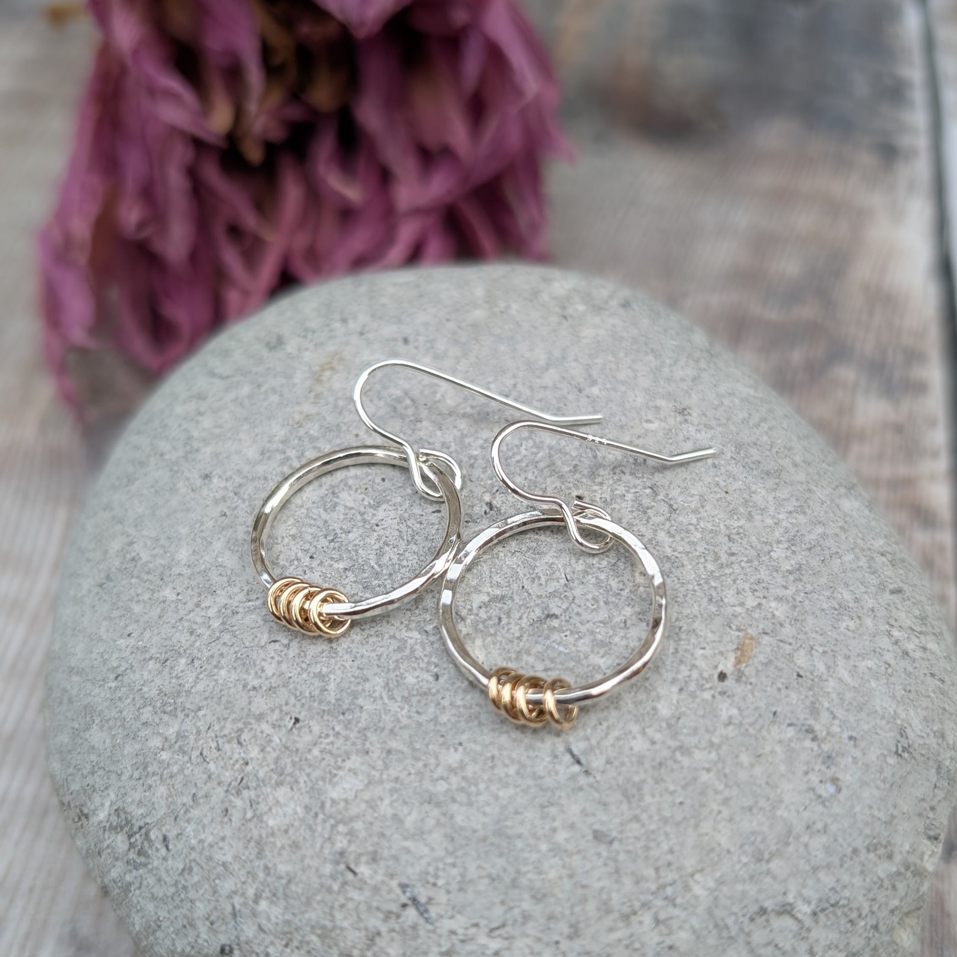 The silver circle earrings with gold loops resting on a smooth grey stone. The earrings overlap slightly, displaying their delicate yet sturdy design. Dried flowers in the background create a soft, natural setting.