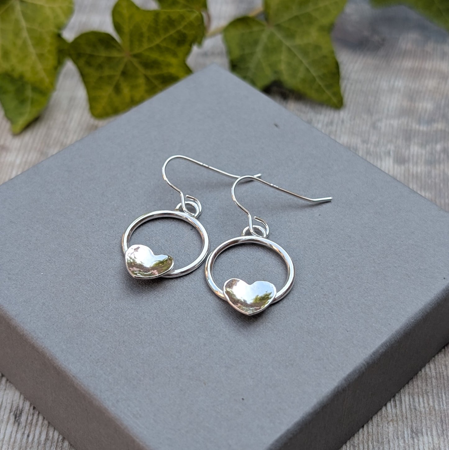 Circle earring with hammered hearts handmade from recycled Sterling Silver, photographed on a grey jewellery box showing the polished silver circles and hammered heart details.