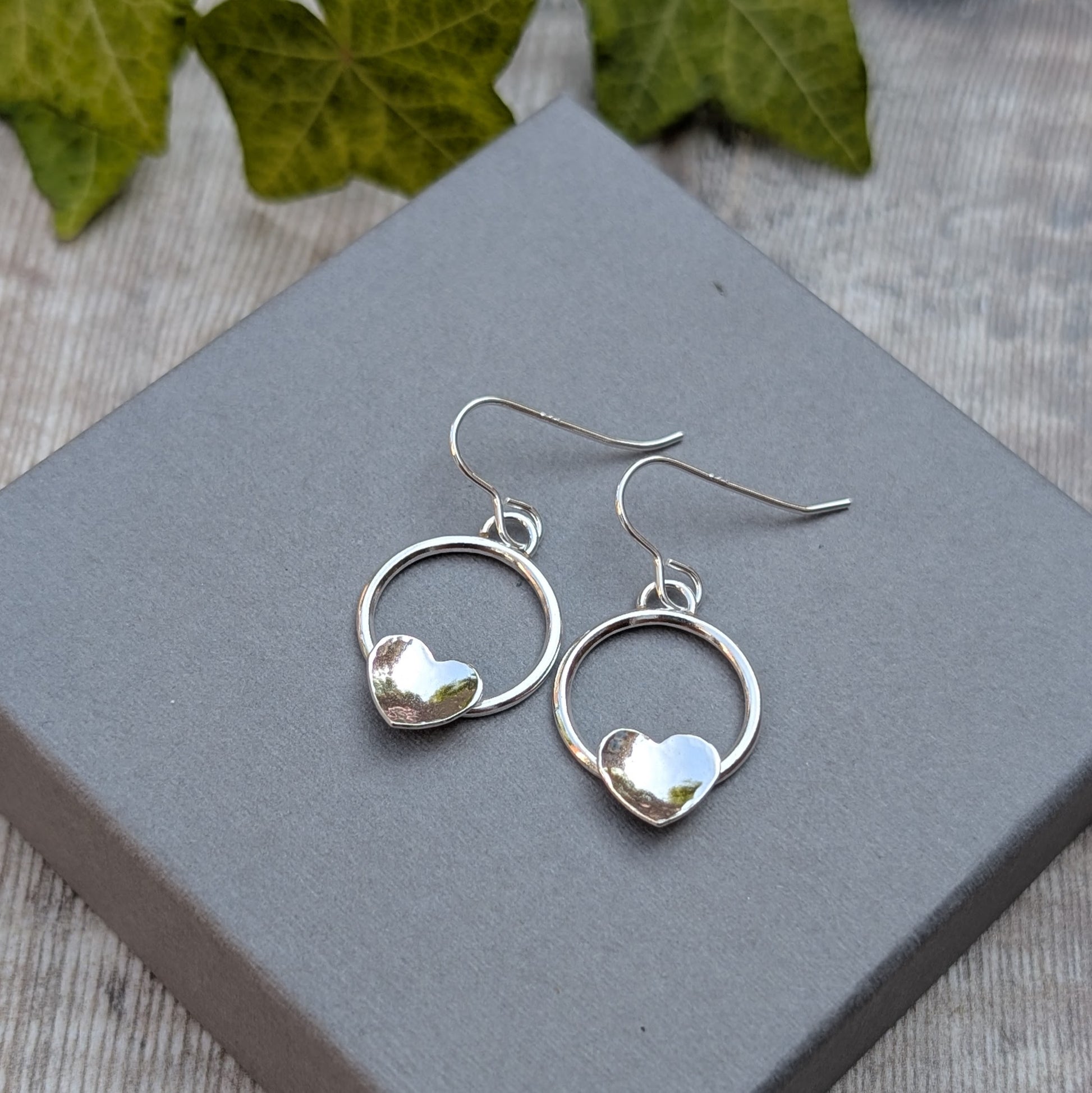 Circle earring with hammered hearts handmade from recycled Sterling Silver, displayed on a grey jewellery box with green ivy leaves in the background.