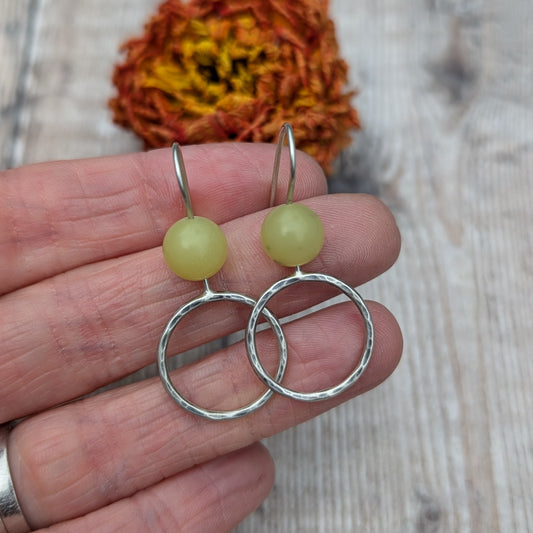 A close-up view of a pair of handmade earrings featuring yellow jade beads and open, hammered silver circles. The earrings are held between fingers against a natural wood background with a dried orange flower in the distance.