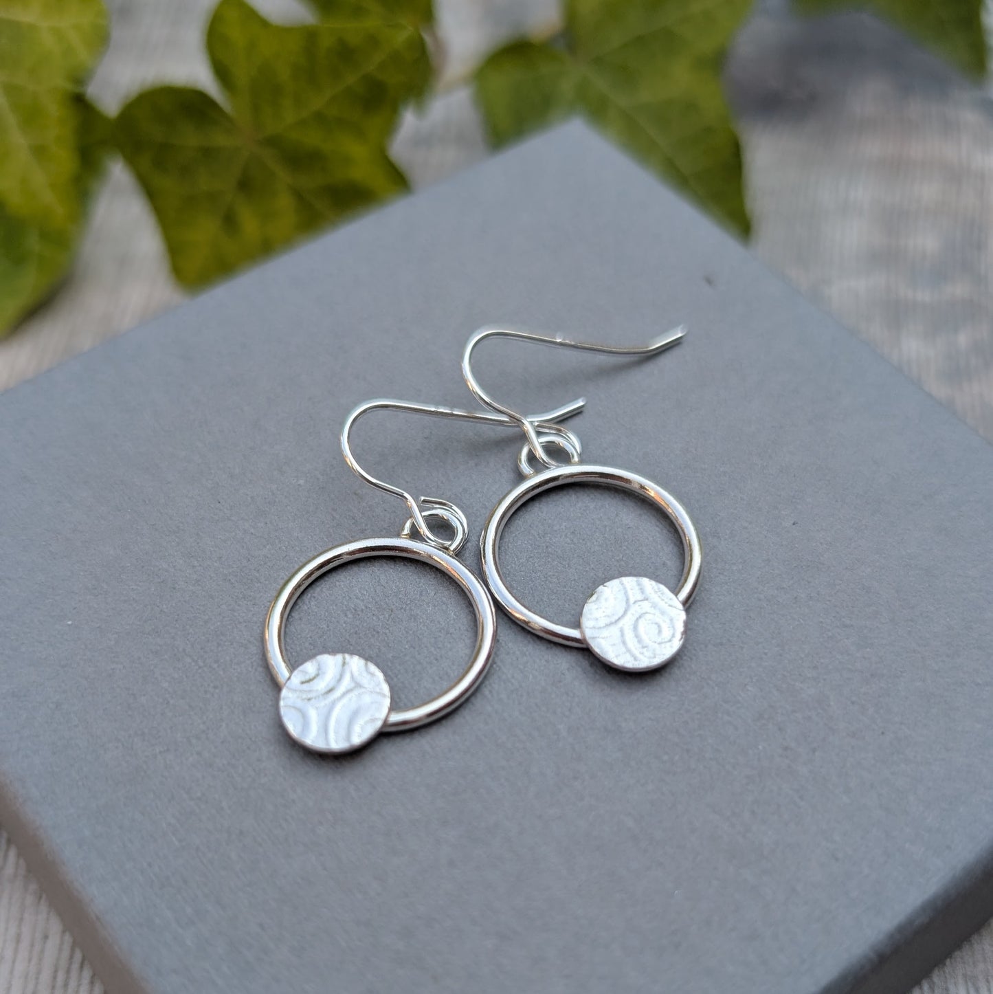Pair of circle earring with textured disc handmade from recycled Sterling Silver displayed on a grey jewellery box with a soft natural background.
