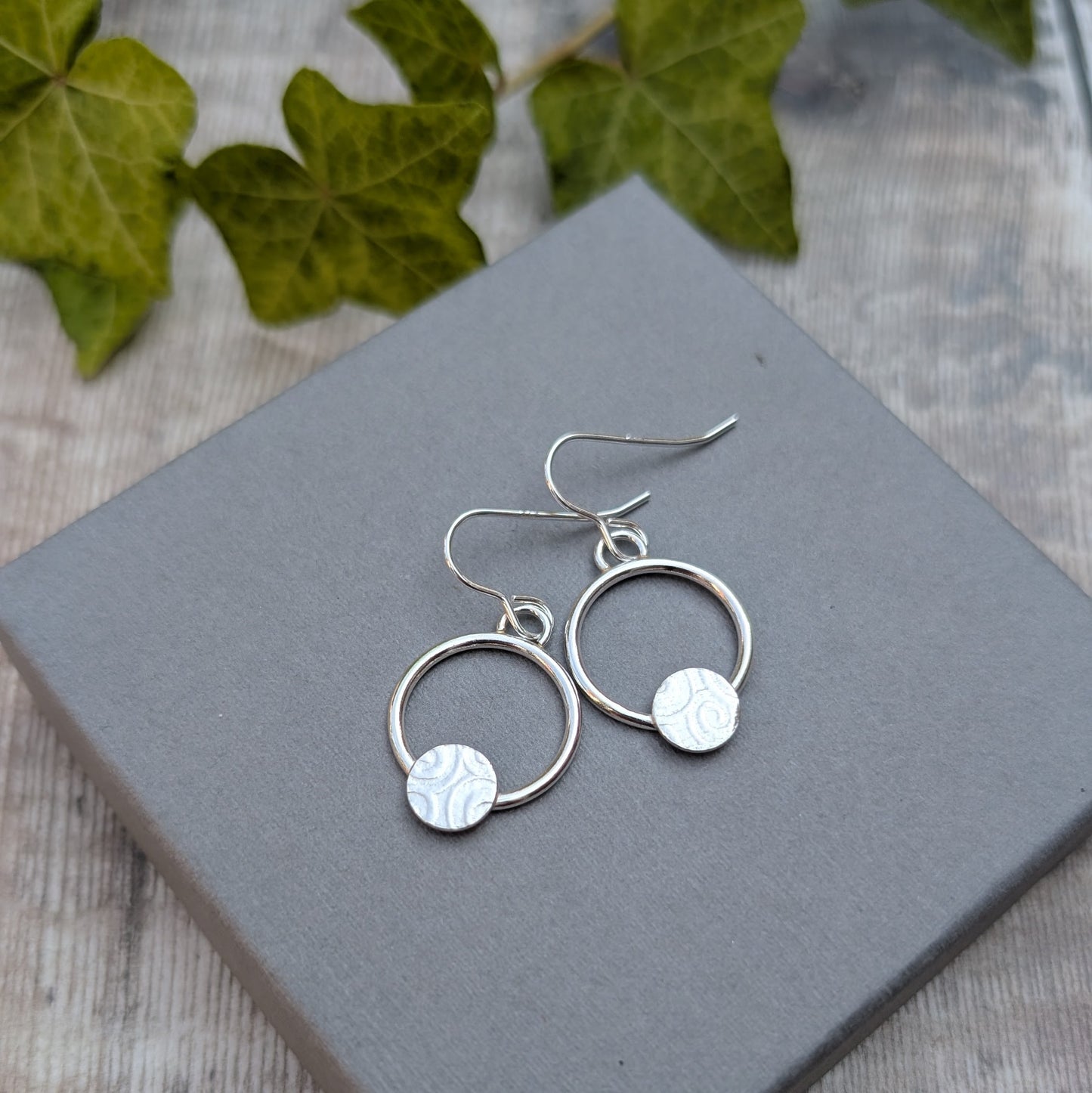 Pair of circle earring with textured disc handmade from recycled Sterling Silver on a grey jewellery box with ivy in the background.
