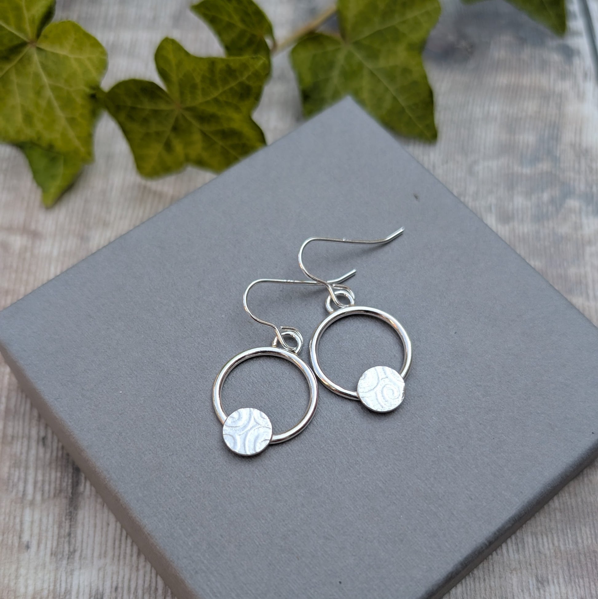 Pair of circle earring with textured disc handmade from recycled Sterling Silver on a grey jewellery box with ivy in the background.