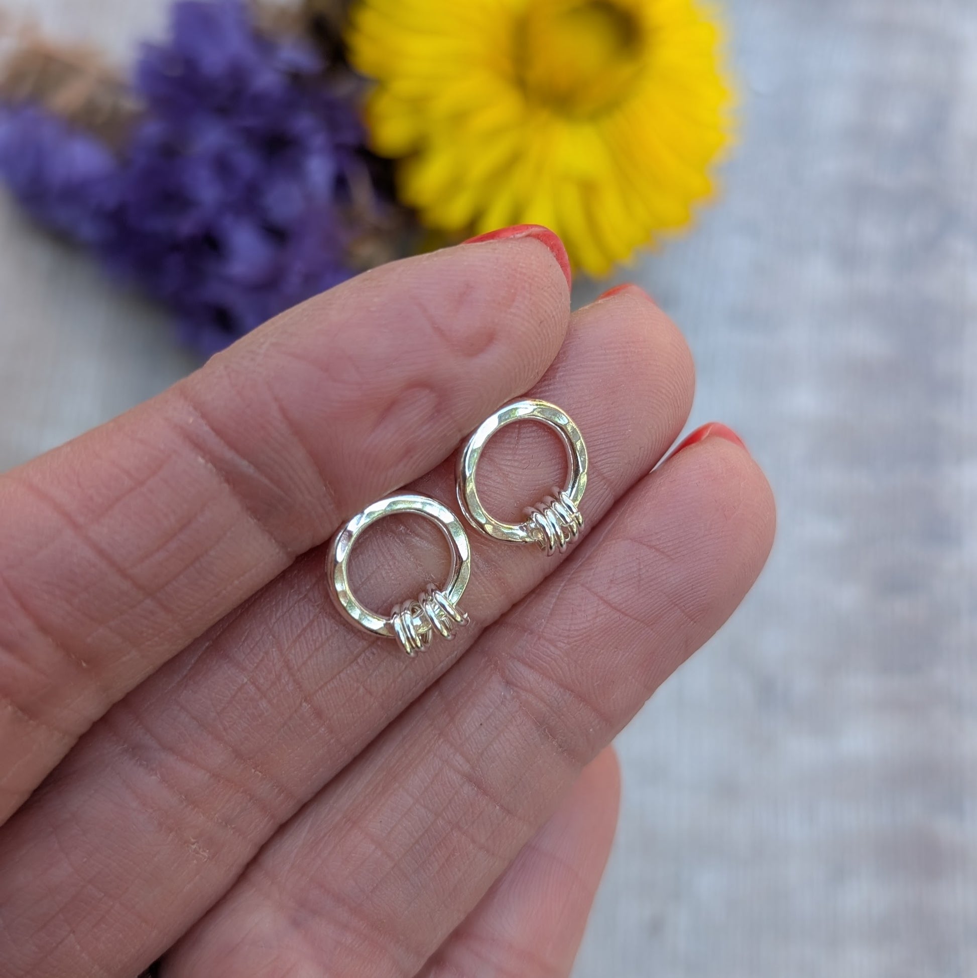 A hand holding a pair of silver circle stud earrings with a hammered finish. Each earring has three tiny silver loops attached at the bottom. A blurred background of yellow and purple dried flowers adds a soft, colourful backdrop.