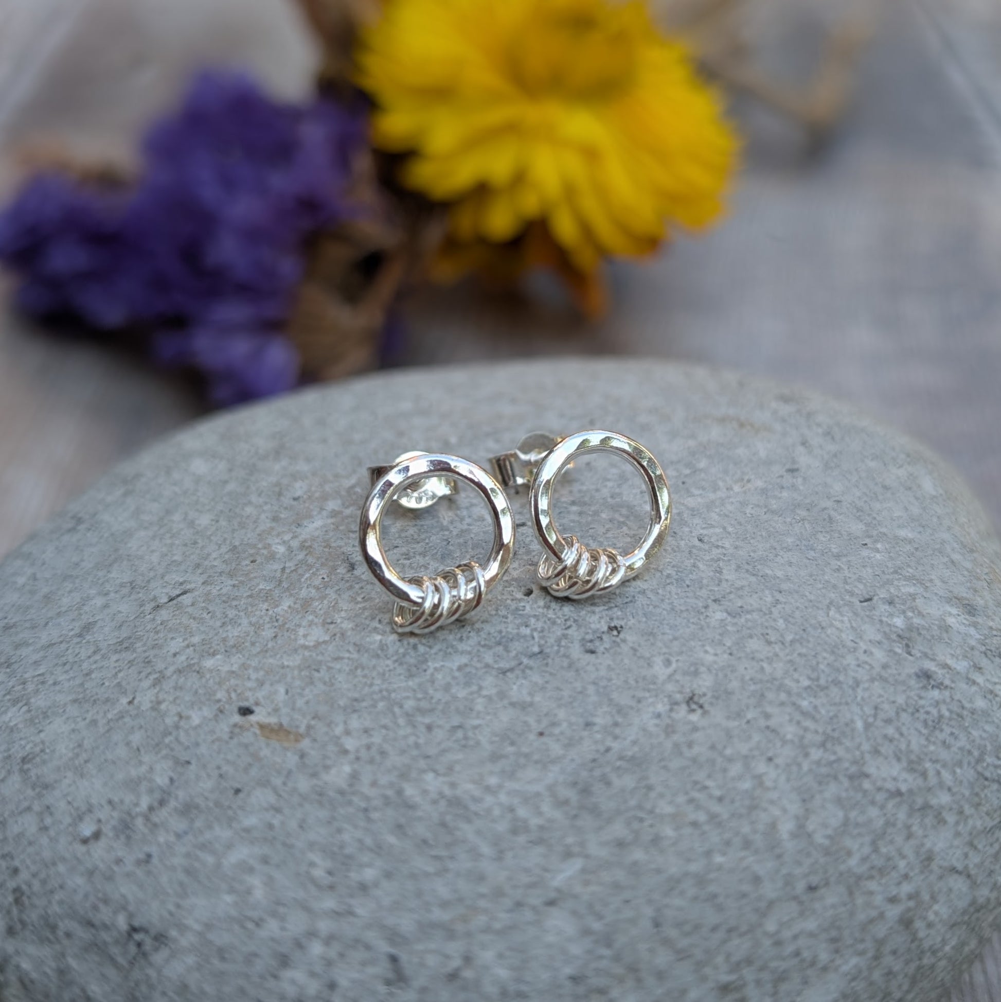 A pair of handmade silver circle stud earrings resting on a smooth grey stone. Each earring features a hammered silver circle with three small silver loops at the bottom. A blurred background of purple and yellow dried flowers adds a natural touch.