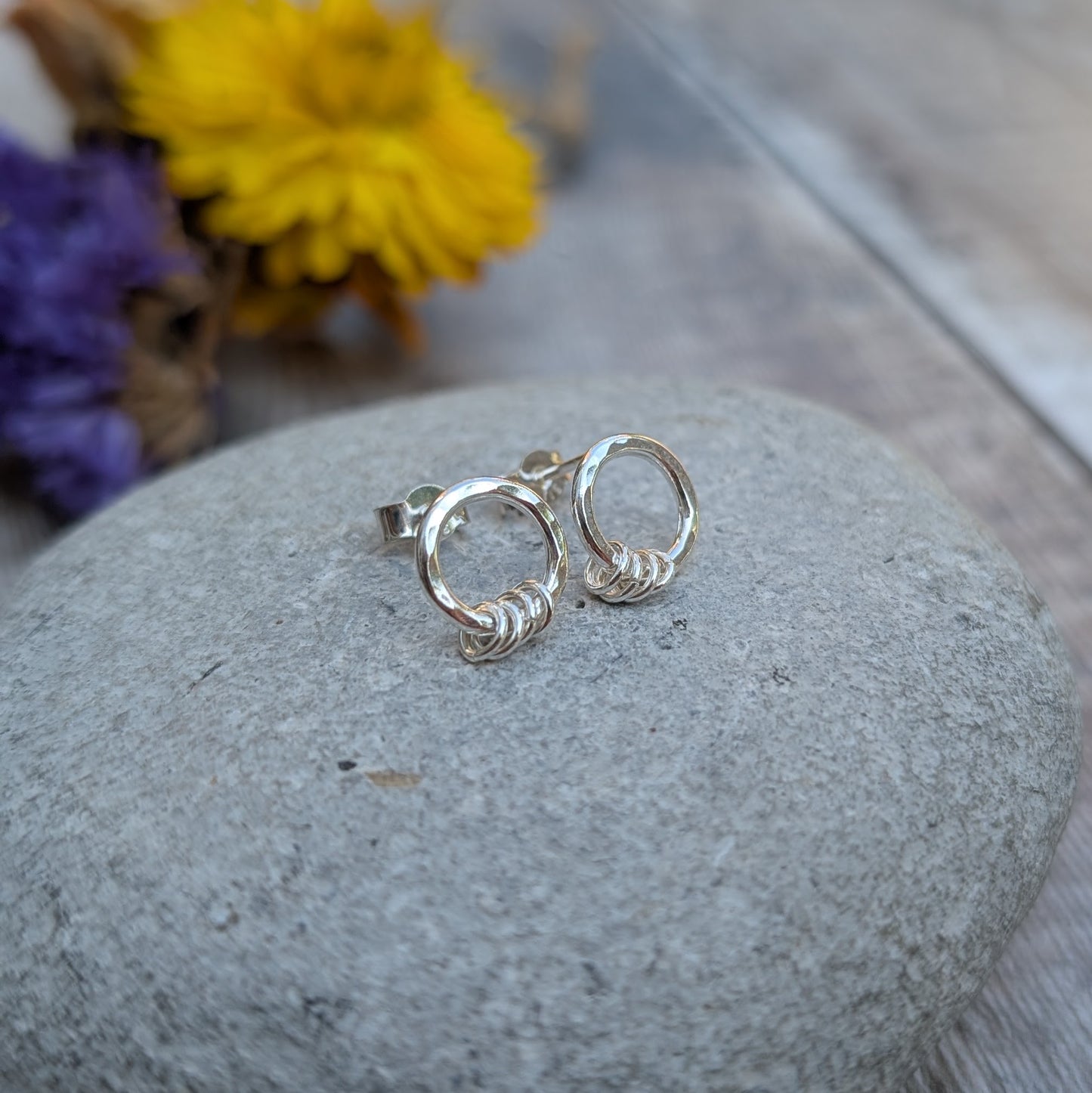 A pair of handmade silver circle stud earrings resting on a smooth grey stone. Each earring features a hammered silver circle with three small silver loops at the bottom. A blurred background of purple and yellow dried flowers adds a natural touch.