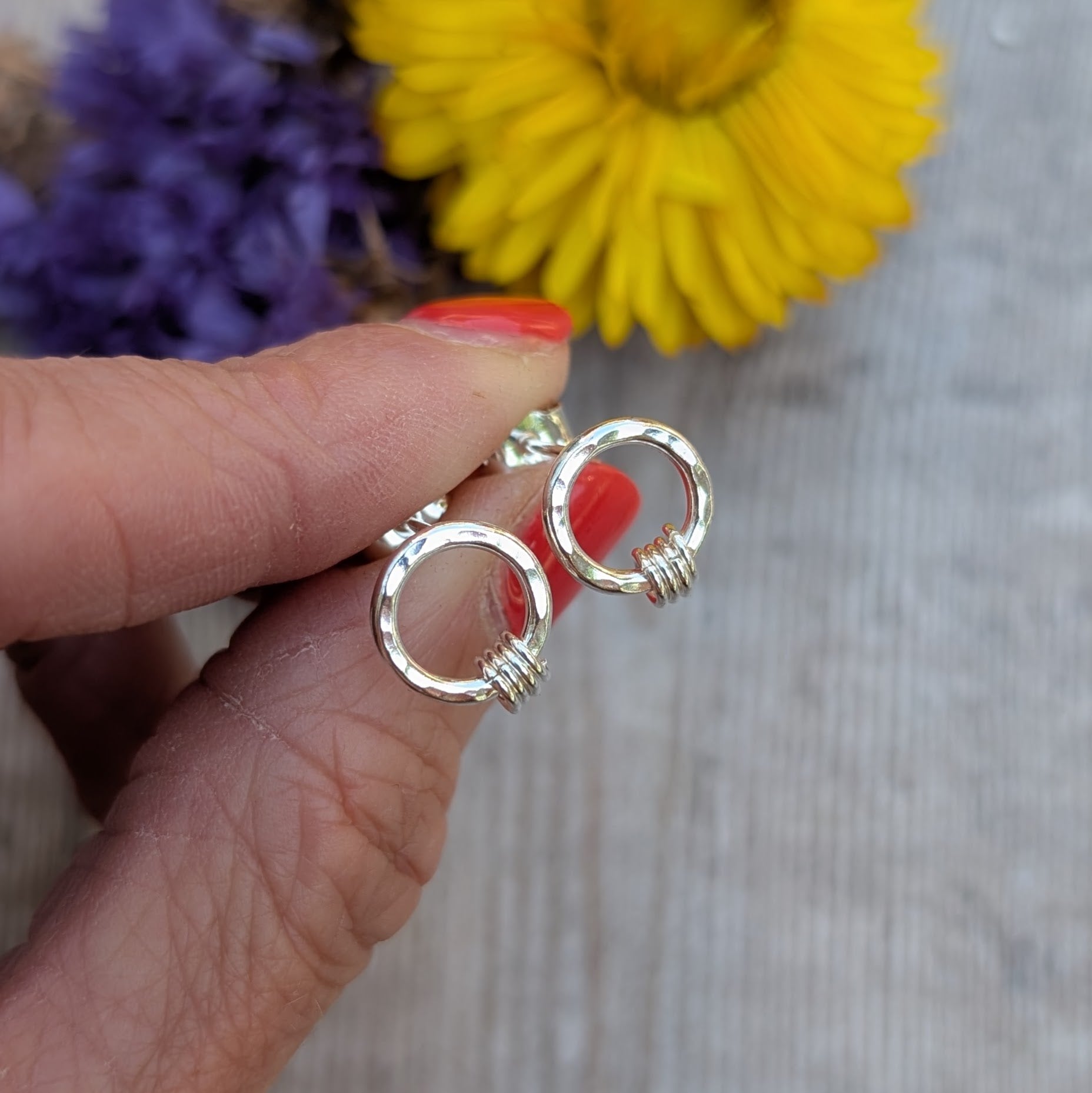 A hand holding a pair of silver circle stud earrings with a hammered finish. Each earring has three tiny silver loops attached at the bottom. A blurred background of yellow and purple dried flowers adds a soft, colourful backdrop.