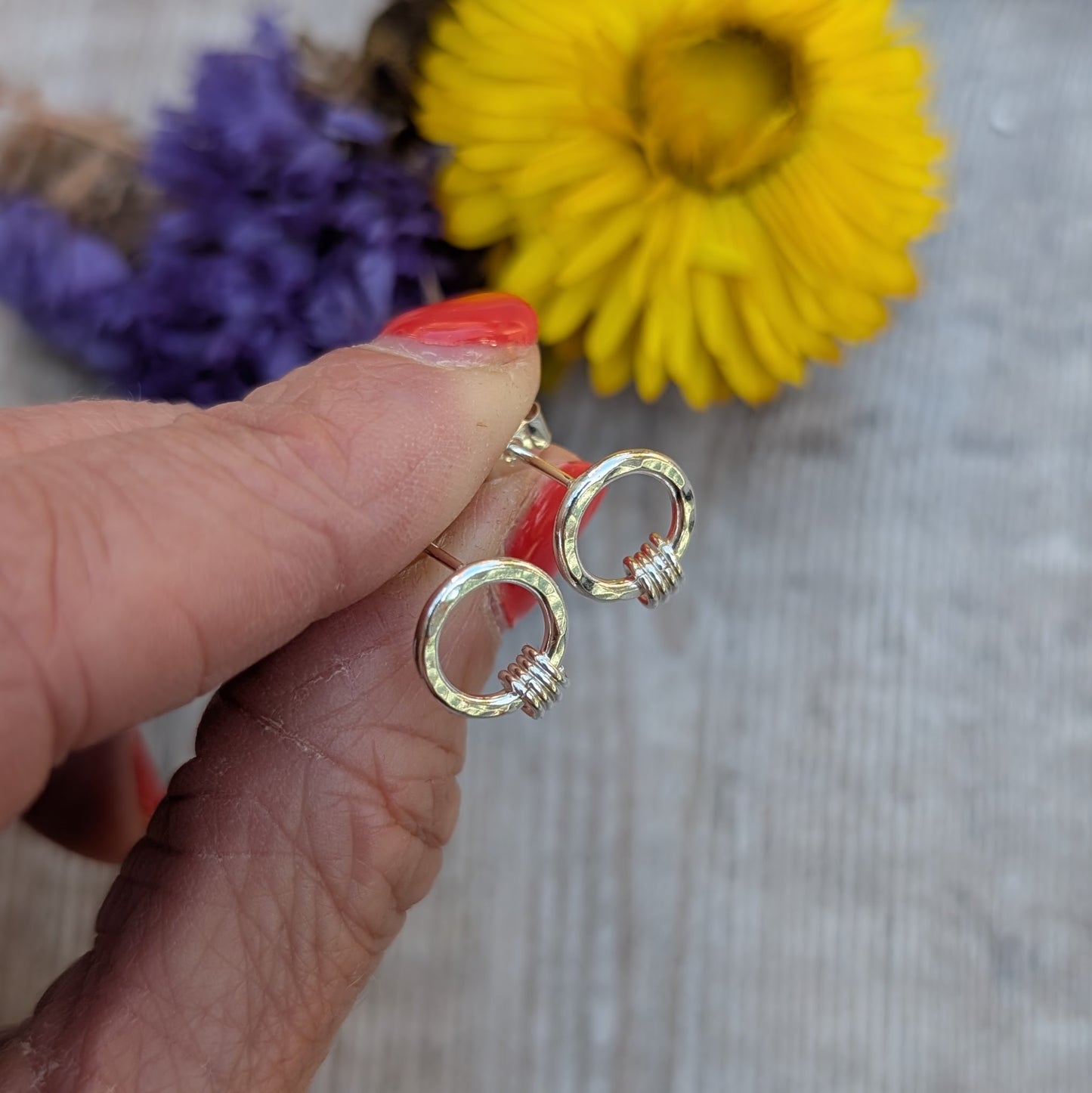 A hand holding a pair of silver circle stud earrings with a hammered finish. Each earring has three tiny silver loops attached at the bottom. A blurred background of yellow and purple dried flowers adds a soft, colourful backdrop.