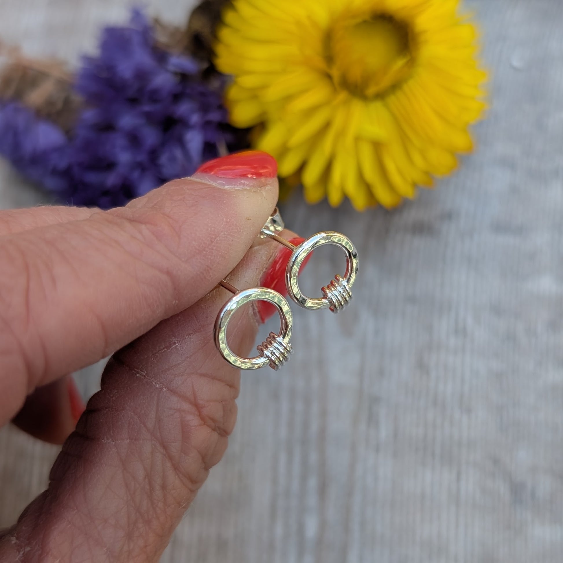 A hand holding a pair of silver circle stud earrings with a hammered finish. Each earring has three tiny silver loops attached at the bottom. A blurred background of yellow and purple dried flowers adds a soft, colourful backdrop.