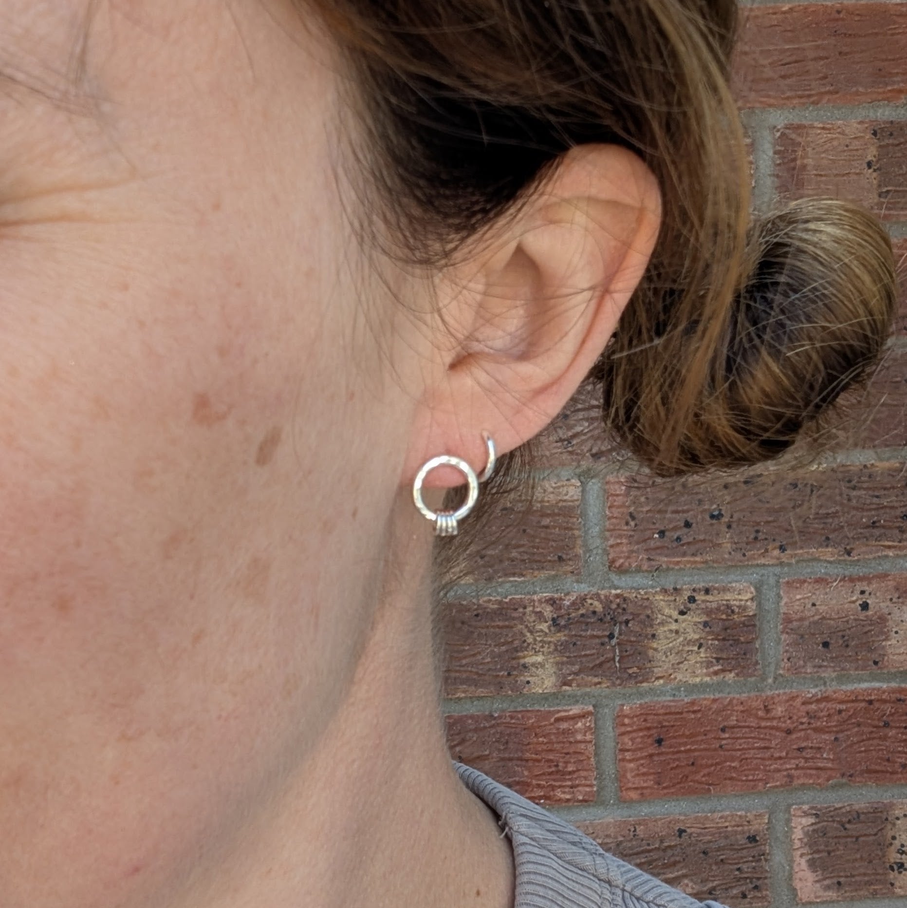  close-up of a person wearing one of the silver circle stud earrings. The hammered silver circle sits neatly on the earlobe with three small silver loops hanging at the bottom. The person’s hair is tied back, and a brick wall is visible in the background.