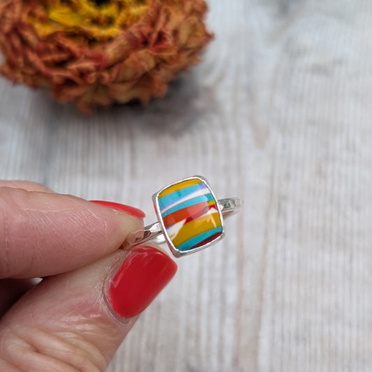 Close-up of a handmade silver ring with a colourful rectangular Surfite stone held between fingers with red nail polish.