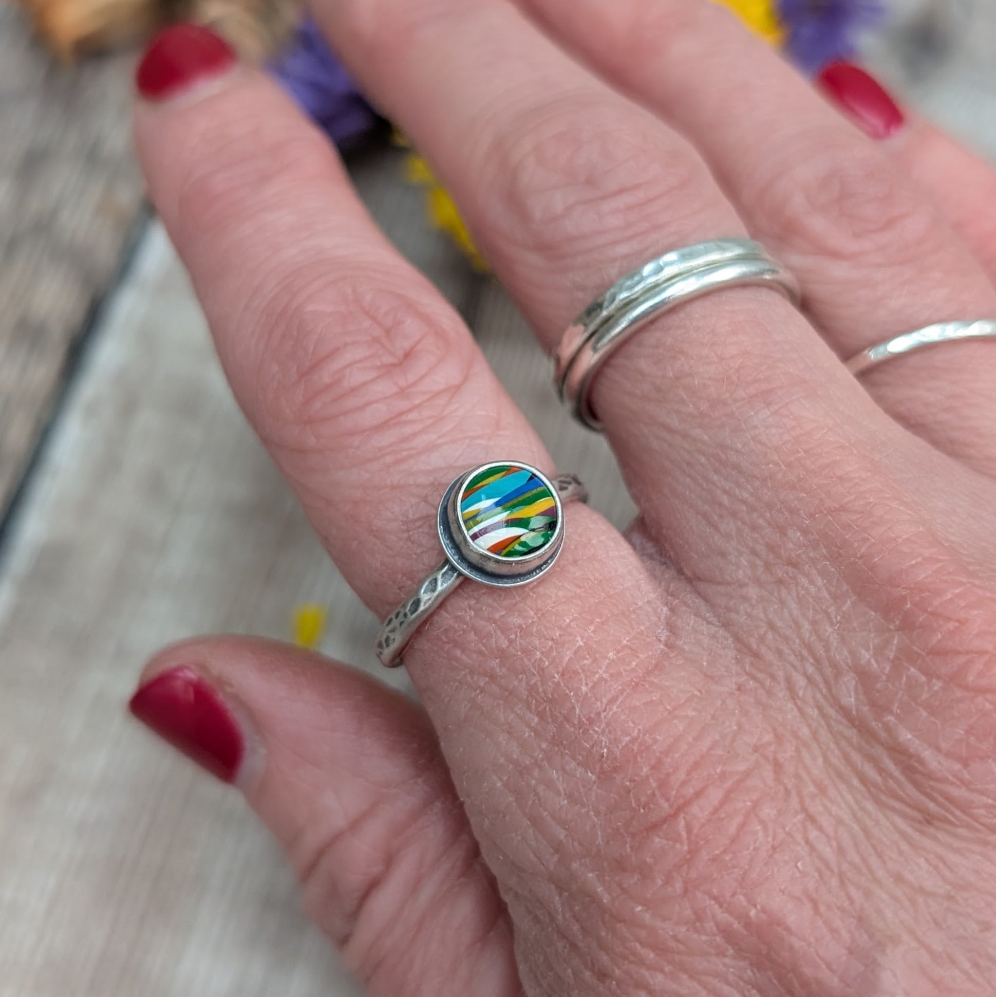 Round surfite stone set in oxidised silver bezel surround. Ring band has textured hammered finish, enhanced by oxidisation. Surfite has uneven horizontal stripes and flecks of green, yellow, blue and white.