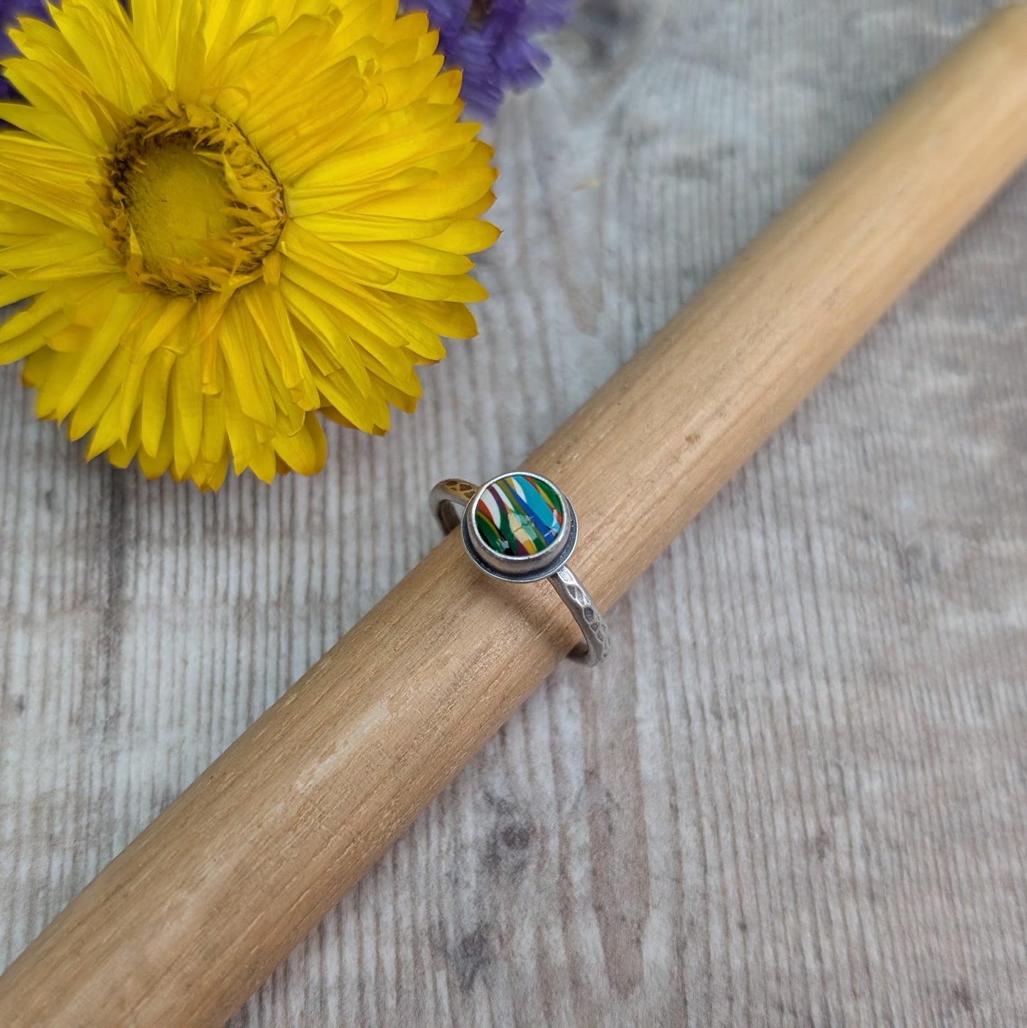 Round surfite stone set in oxidised silver bezel surround. Ring band has textured hammered finish, enhanced by oxidisation. Surfite has uneven horizontal stripes and flecks of green, yellow, blue and white.