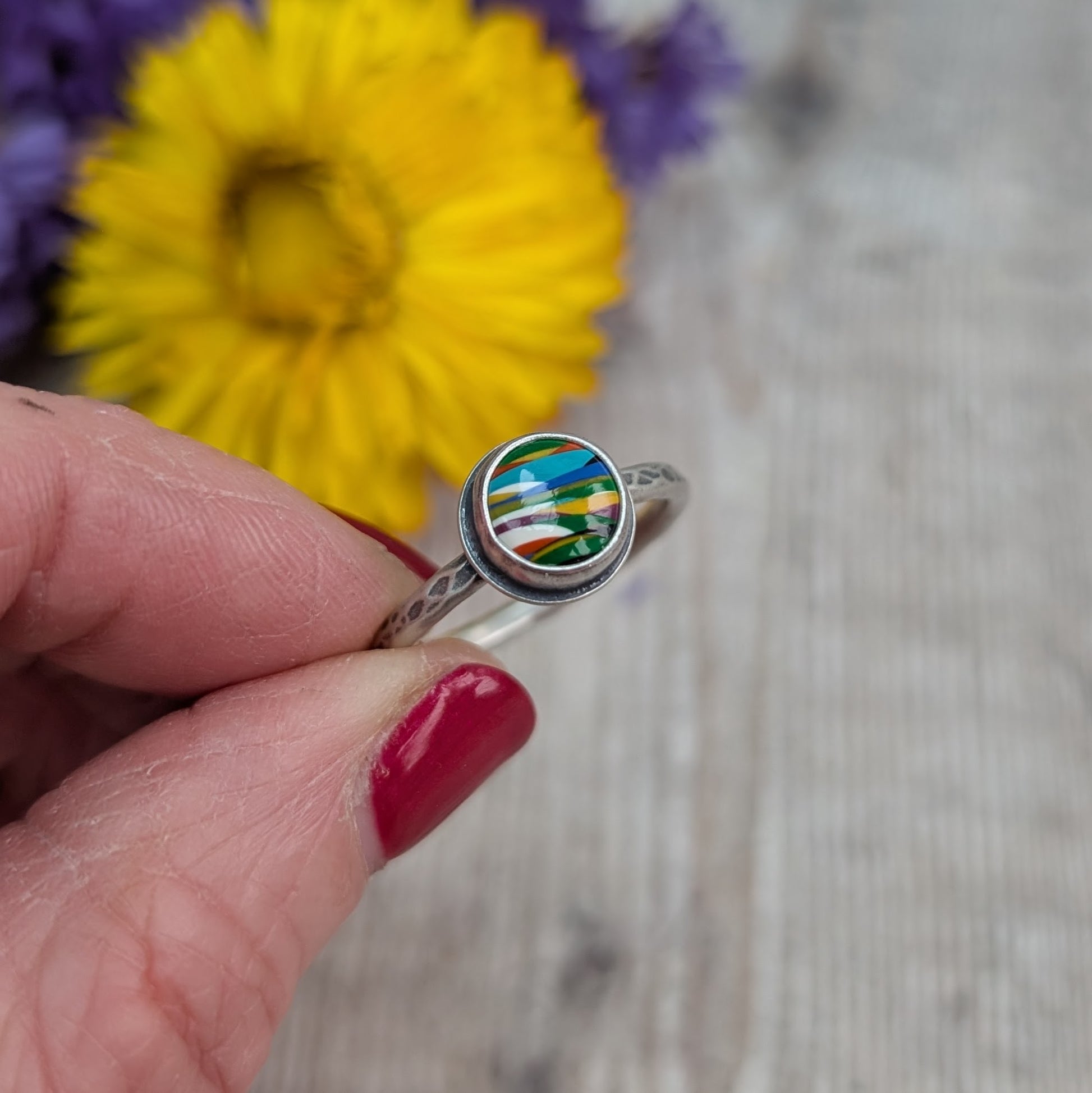 Round surfite stone set in oxidised silver bezel surround. Ring band has textured hammered finish, enhanced by oxidisation. Surfite has uneven horizontal stripes and flecks of green, yellow, blue and white.