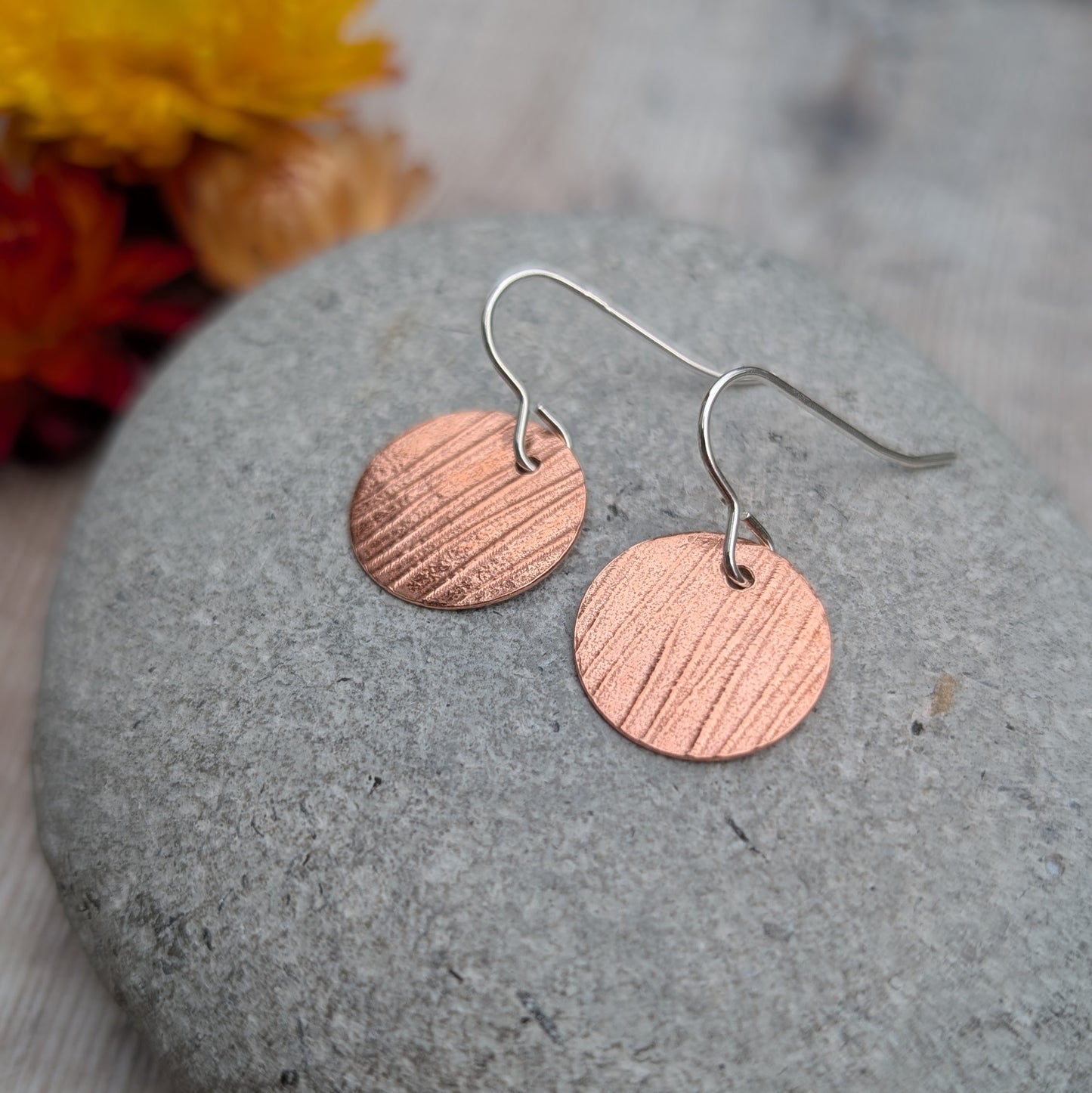 Copper bark pattern disc earrings resting on a smooth grey stone showing the lined bark texture pressed into the copper.