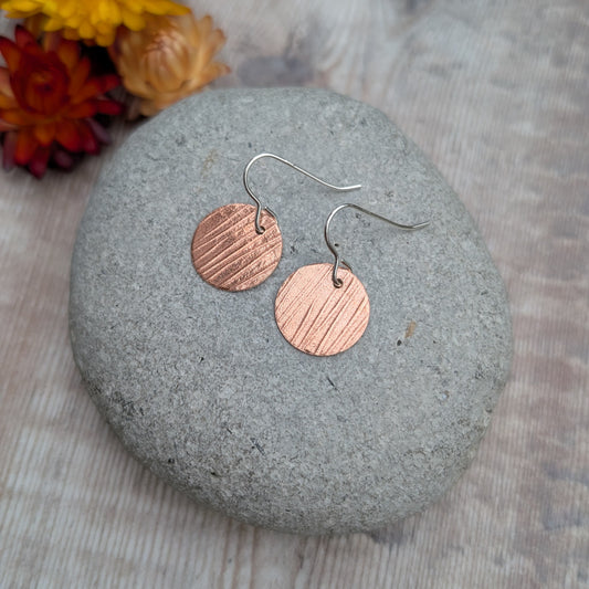 Pair of Copper bark pattern disc earrings displayed on a stone with dried flowers in the background.