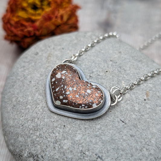 A close-up image of a heart-shaped Copper Firebrick pendant set in recycled sterling silver, featuring warm copper tones with white speckles. The pendant rests on a grey stone background with a dried flower in the background.