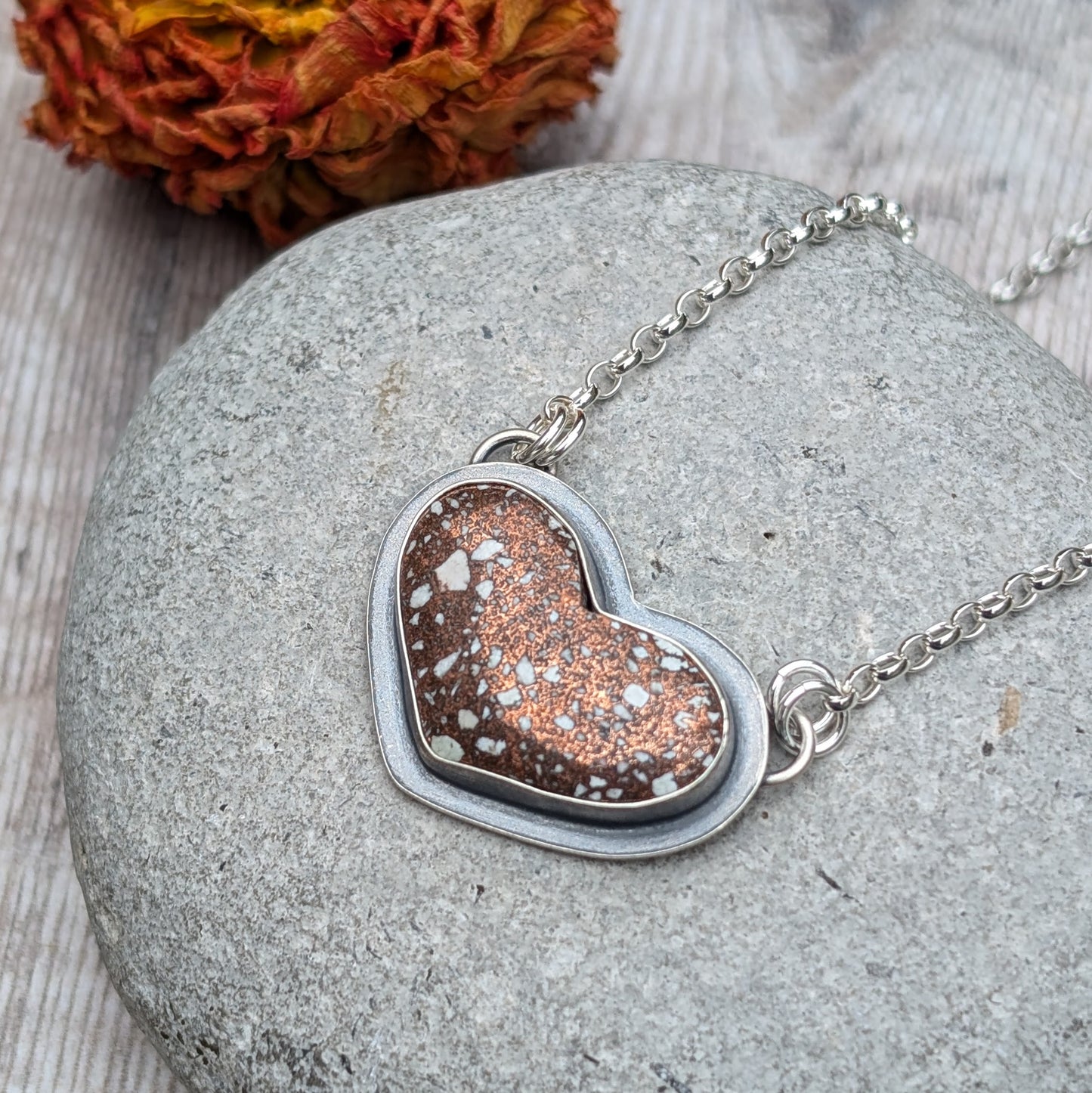 A close-up image of a heart-shaped Copper Firebrick pendant set in recycled sterling silver, featuring warm copper tones with white speckles. The pendant rests on a grey stone background with a dried flower in the background.