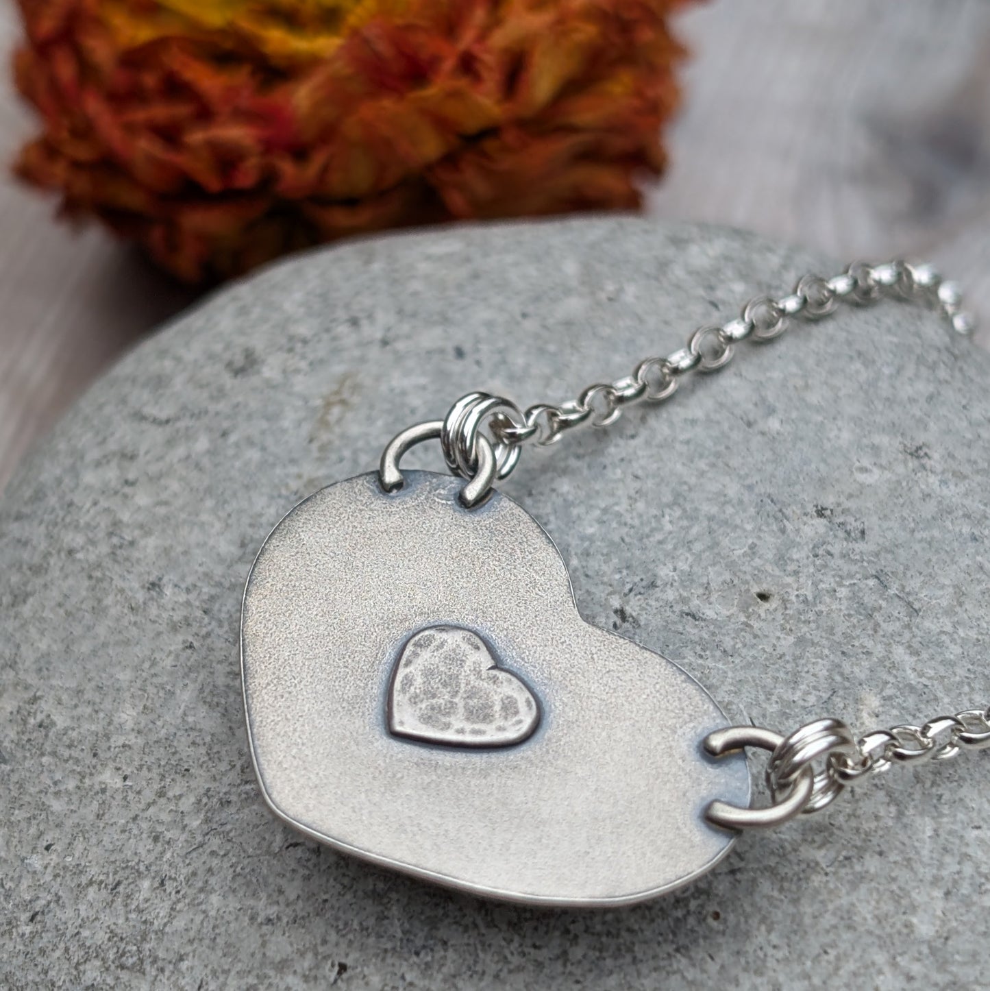 The reverse side of a heart-shaped pendant showing a small silver heart detail set in a textured sterling silver frame. The necklace is placed on a grey stone surface with a dried flower in the background.