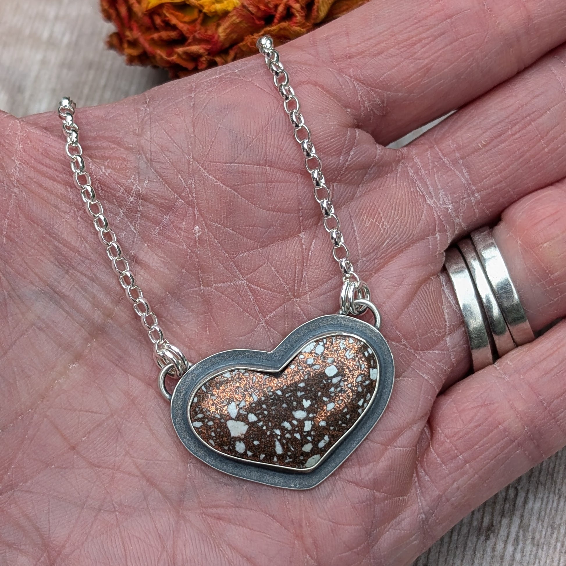 The Copper Firebrick Heart Necklace being held in a hand, showcasing the pendant’s size relative to the hand. The warm copper tones with white speckles are highlighted against the silver frame.