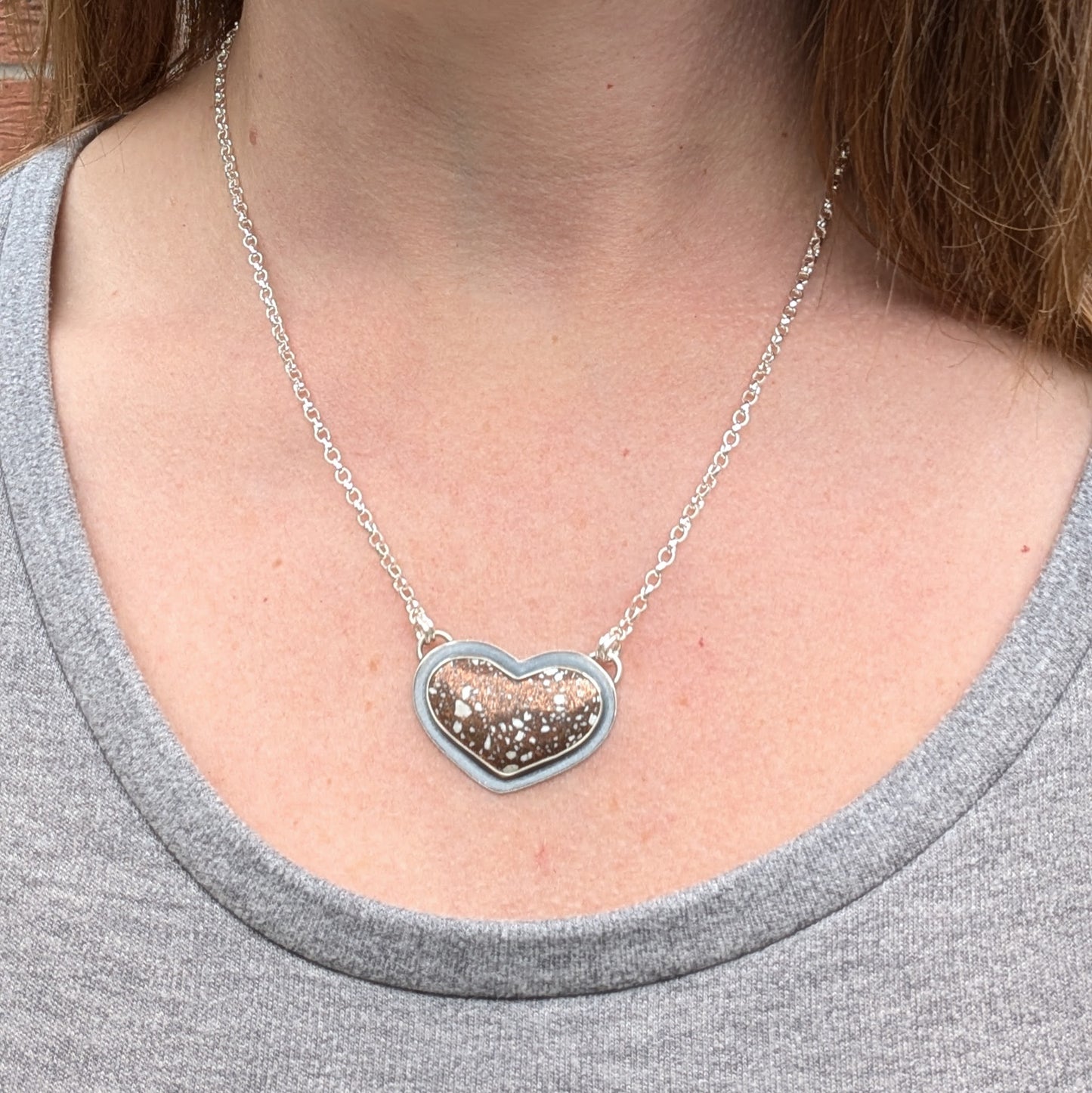 The Copper Firebrick Heart Necklace being worn by a person, showing the pendant resting on a silver chain around the neck. The heart-shaped stone has copper and white speckles, contrasting against a grey shirt.