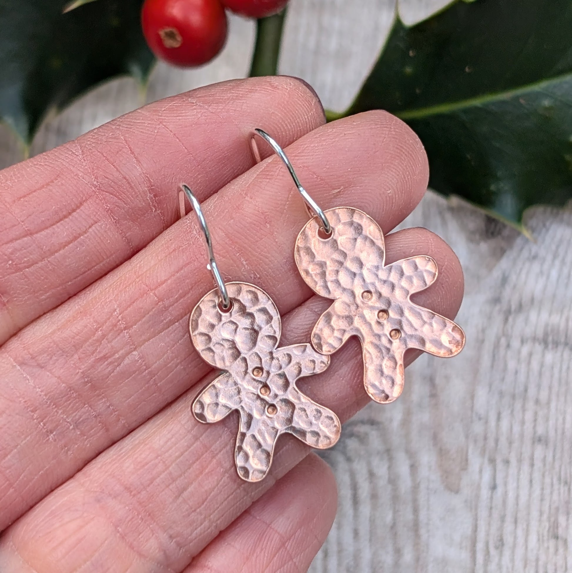 Close-up of two handmade copper gingerbread drop earrings with a hammered texture, being held in a person's hand against a festive background with holly and berries.