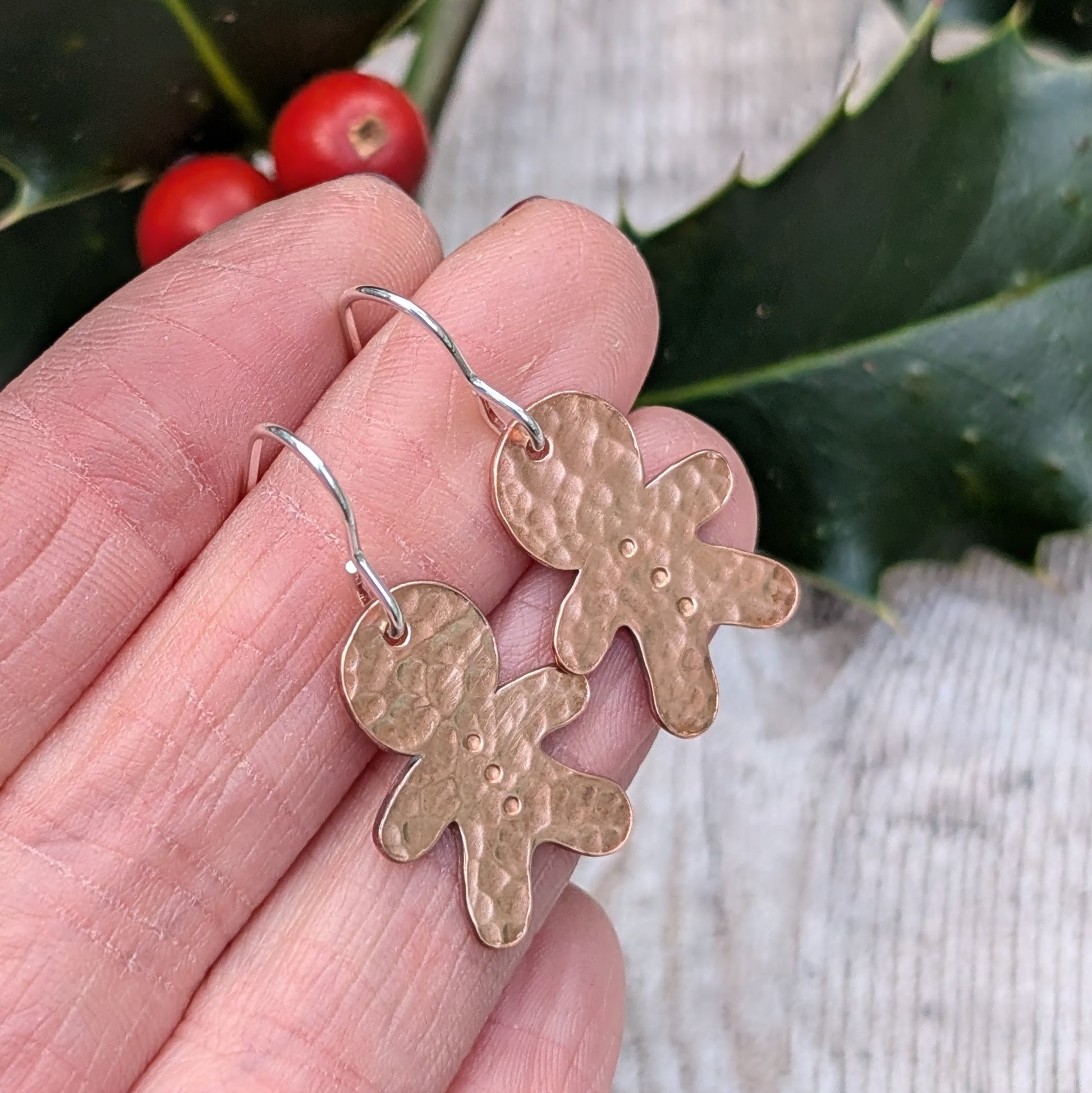 Close-up of two handmade copper gingerbread drop earrings with a hammered texture, being held in a person's hand against a festive background with holly and berries.